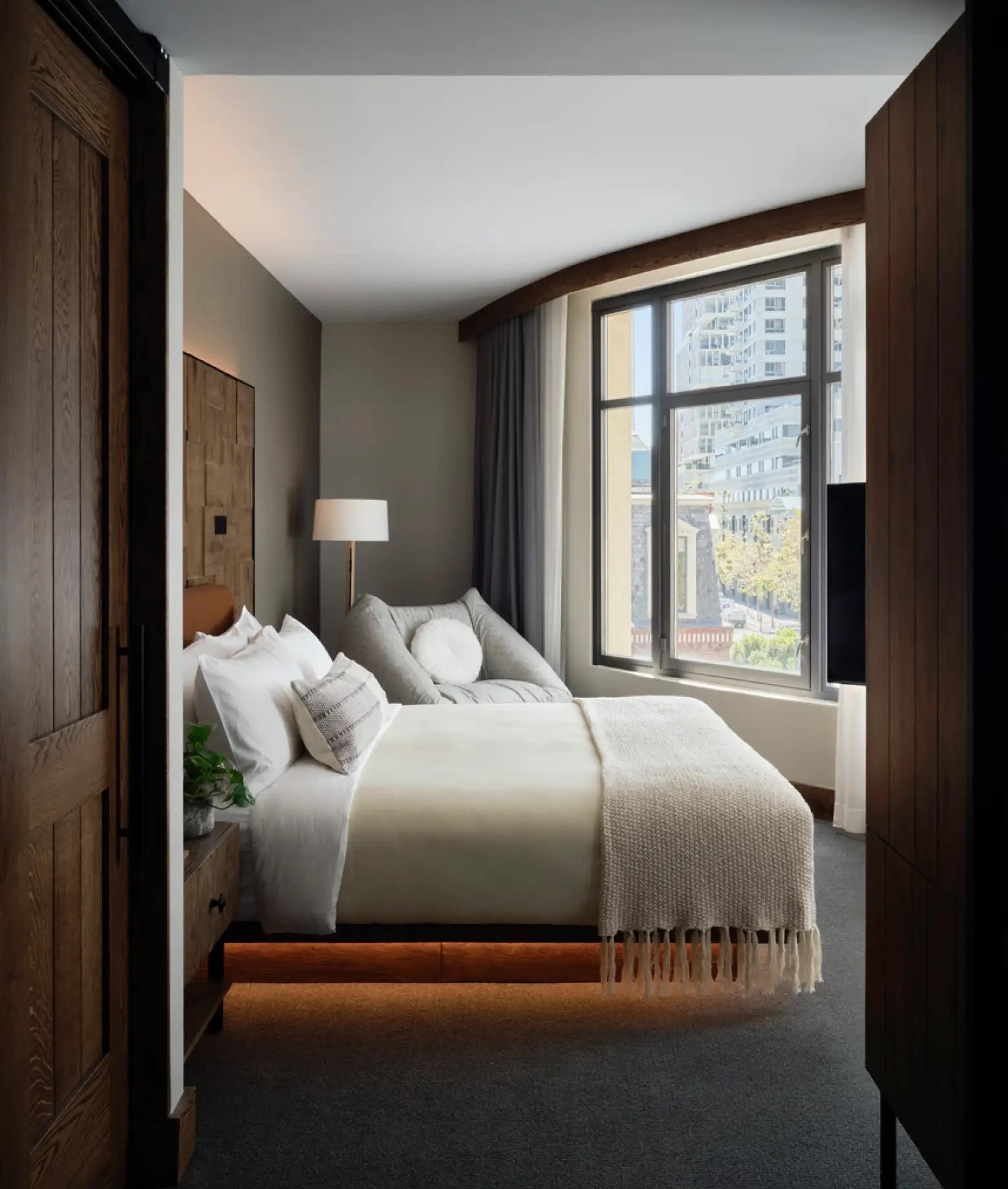 A narrow bedroom seen past a dark wooden door, with a bed under a fringed throw and a window onto a city street.
