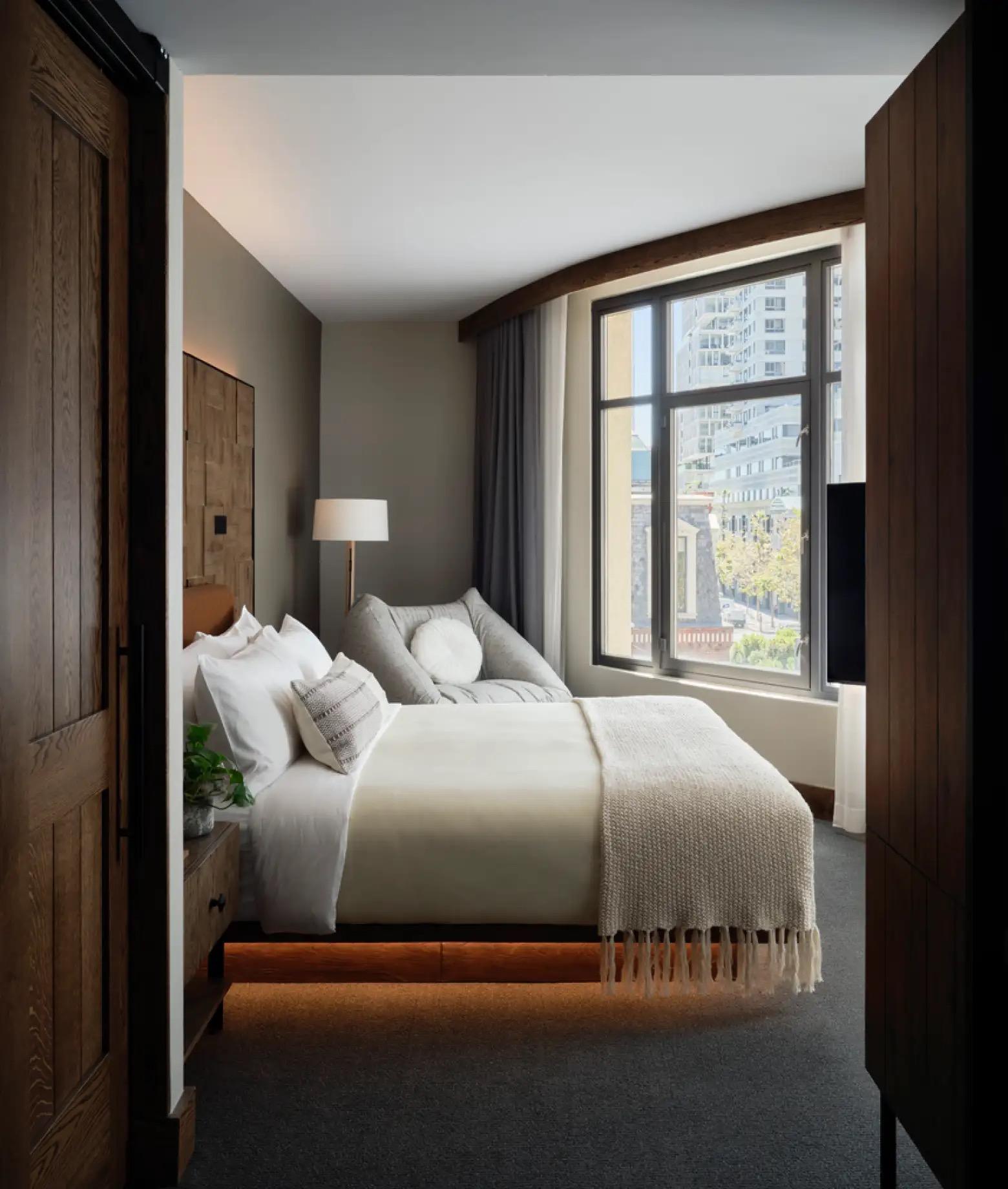 A narrow bedroom seen past a dark wooden door, with a bed under a fringed throw and a window onto a city street.
