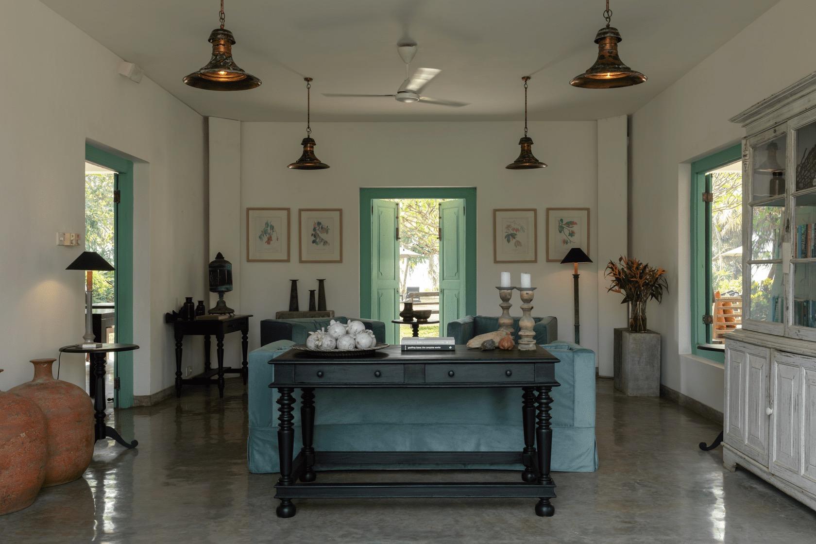 A dark console table holds a dish of shells, a stack of books and two carved stone candlesticks, between two pale blue sofas, with large terracotta urns and pendant lamps to the sides.