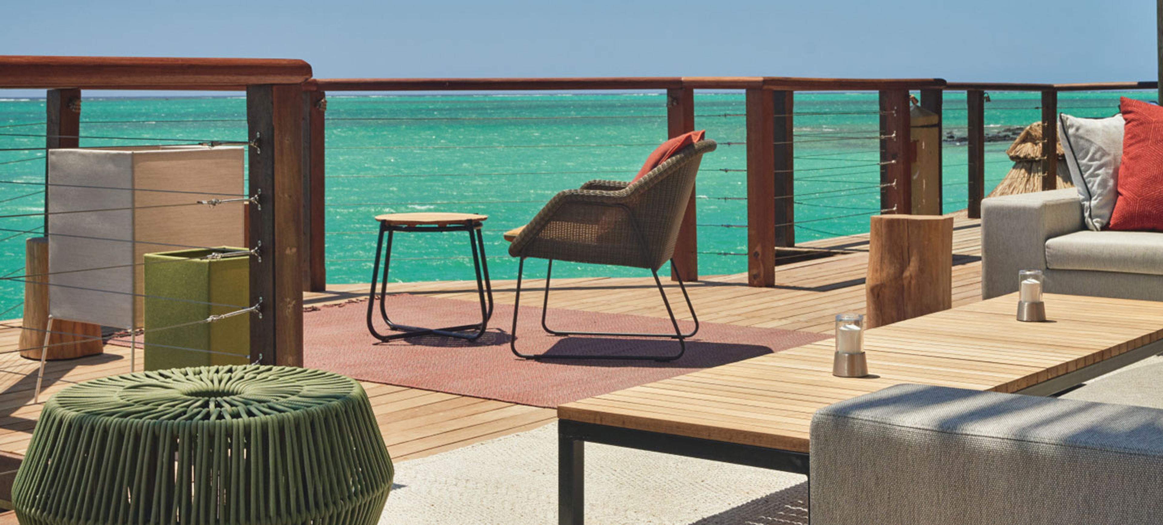 A wooden deck with a cushioned lounge chair, a low table and a sofa against a cable railing, overlooking a turquoise lagoon.