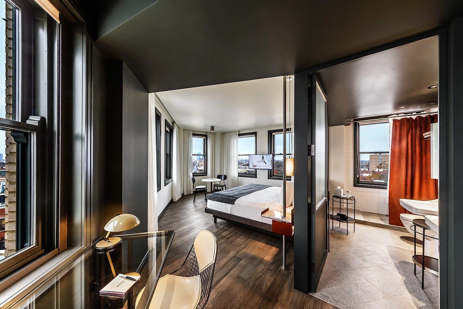 a hotel suite seen from the window corner, with a bed and television along one wall, a desk and chair by the windows, and an open door showing a bathroom with an orange curtain and a view beyond