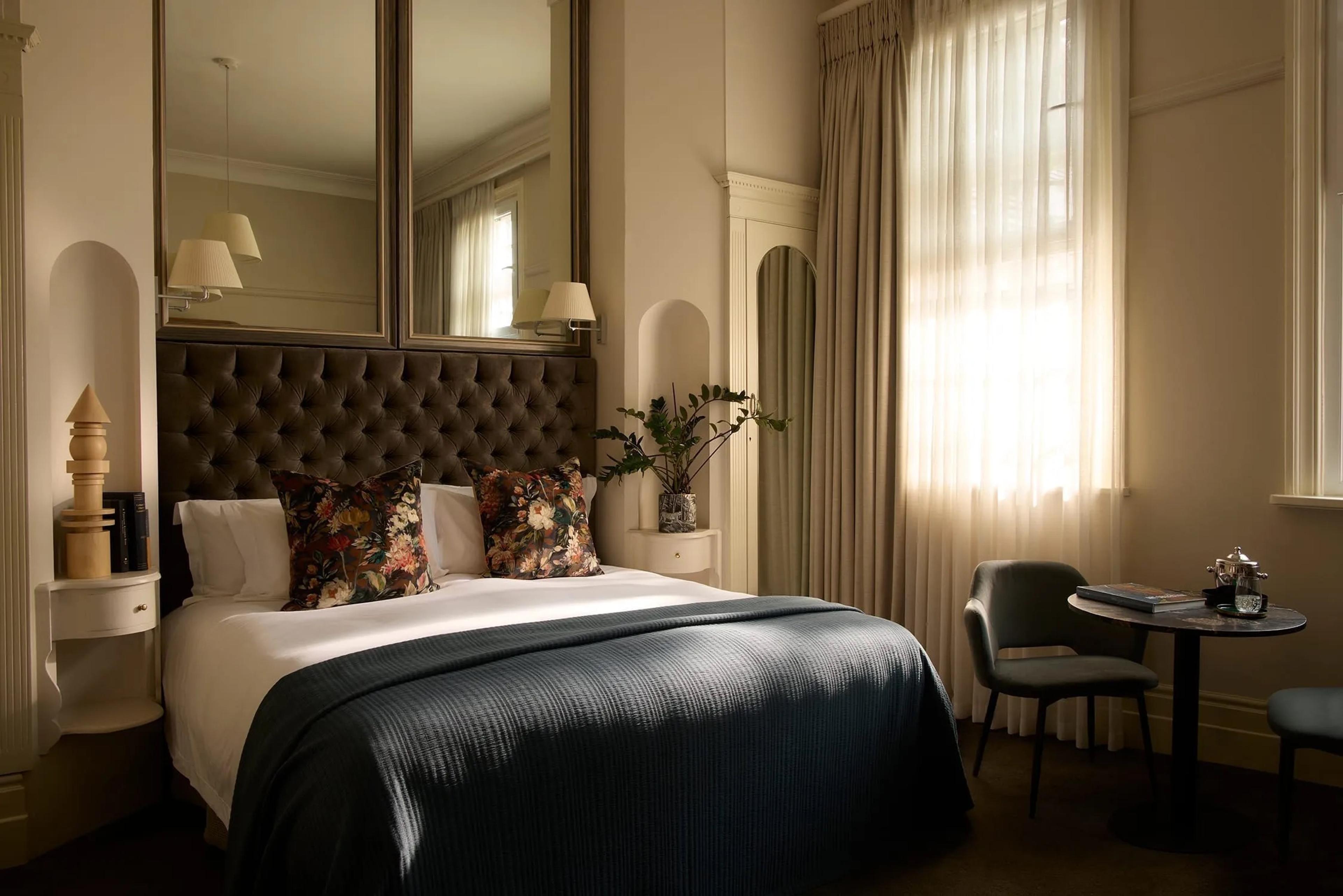 A bedroom with a tufted dark headboard under two tall mirrors, floral cushions, a dark green throw on the bed, and a small round table by a window.