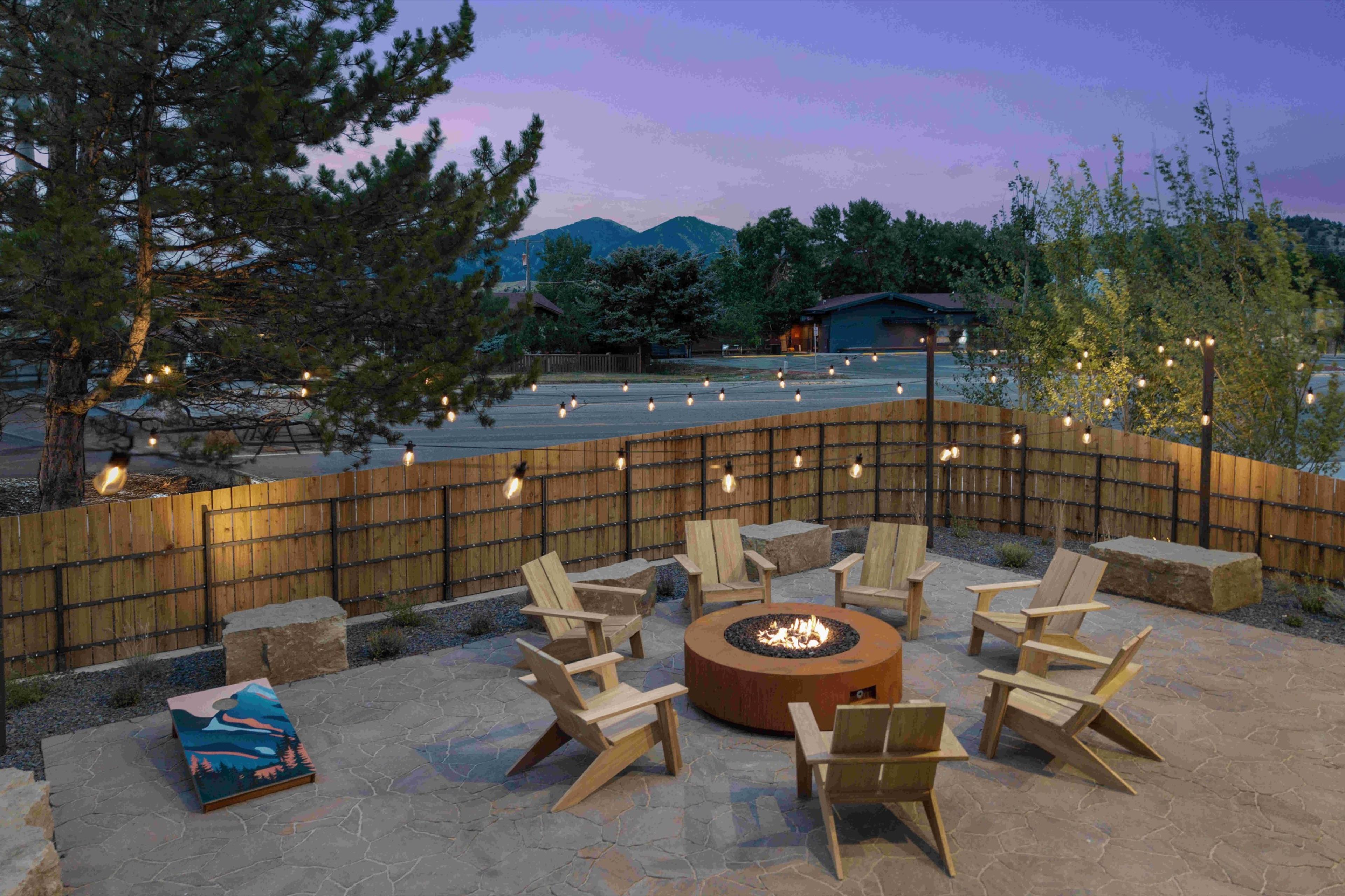 A flagstone patio with wooden chairs around a lit fire pit, enclosed by a strung-light wooden fence, with mountains behind at dusk.