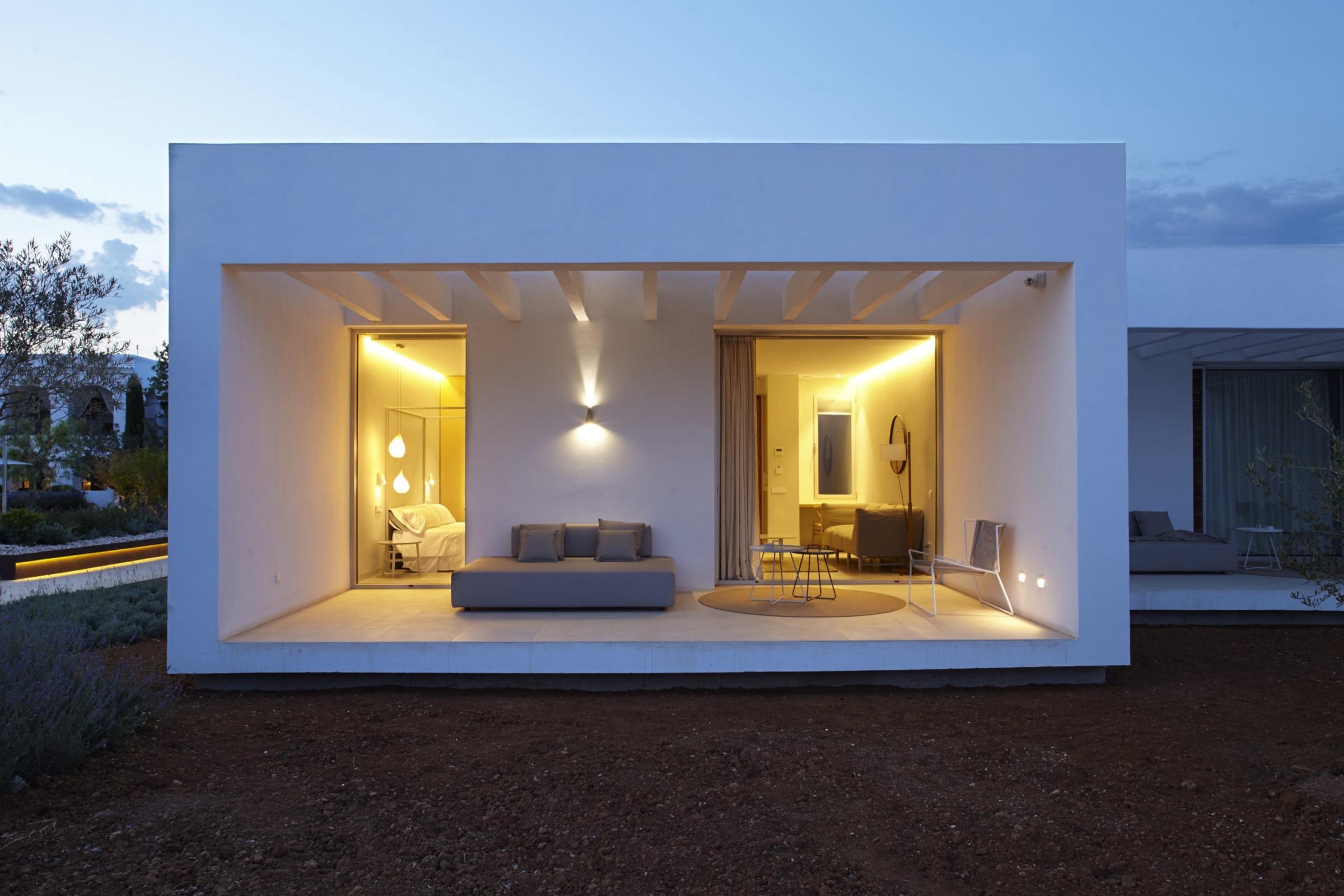 A flat-roofed white building at dusk, its open side showing a lit lounge and bedroom, with a garden in shadow around it.