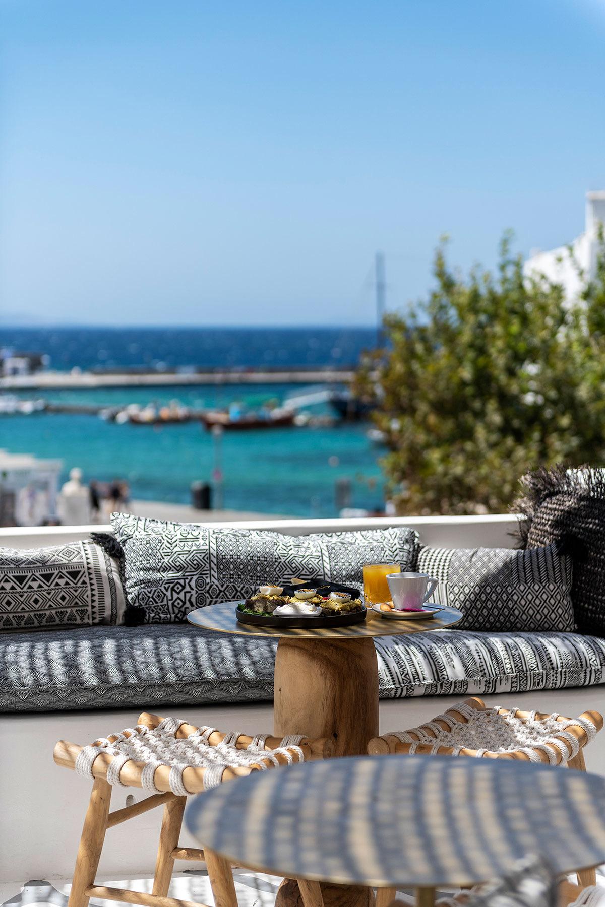 A low table on a sun terrace set with small plates of food, a coffee and a glass of juice, the sea visible beyond.