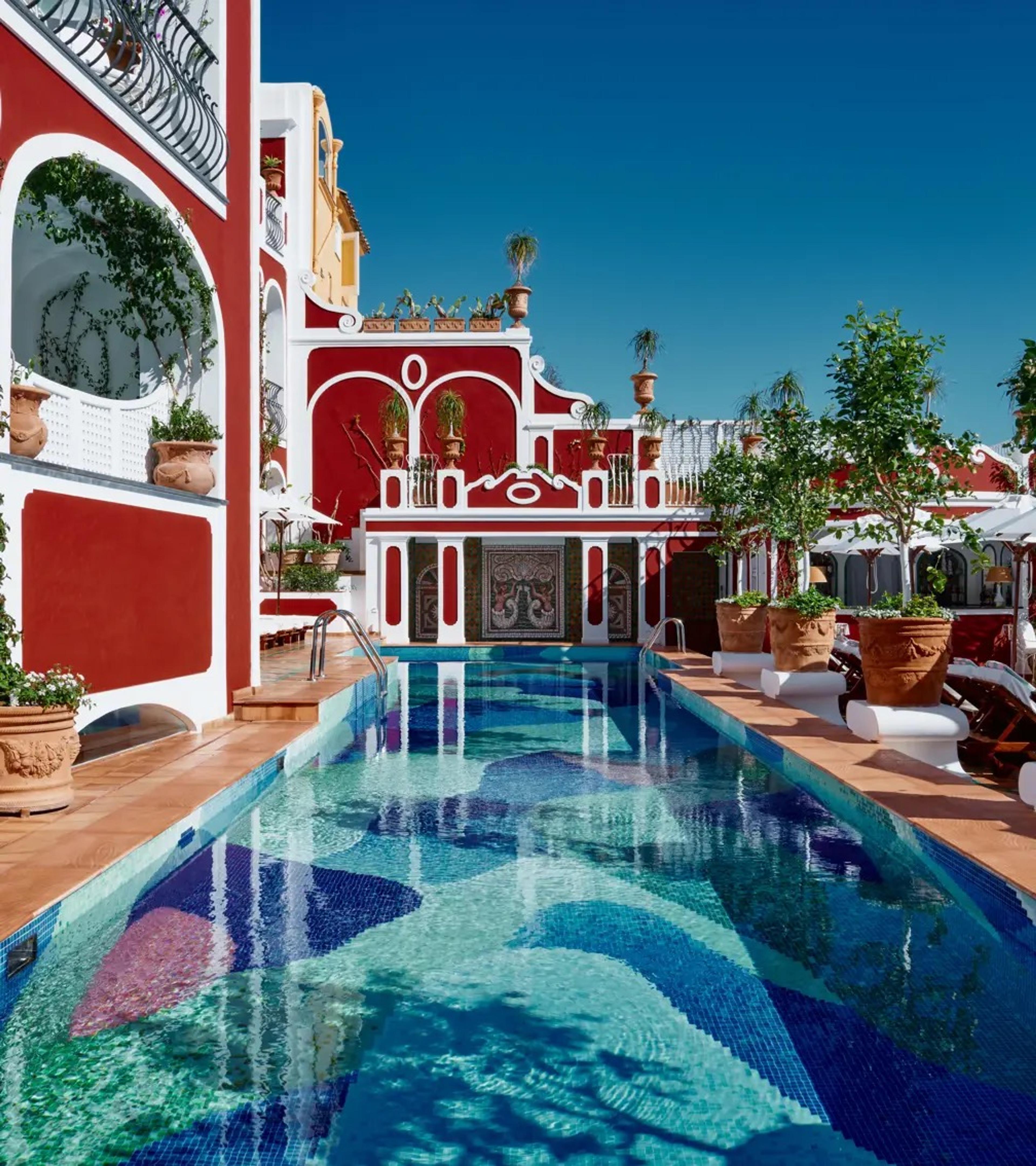 A pool lined in red and blue tiles on a terrace among lemon trees, the hotel's red walls behind.