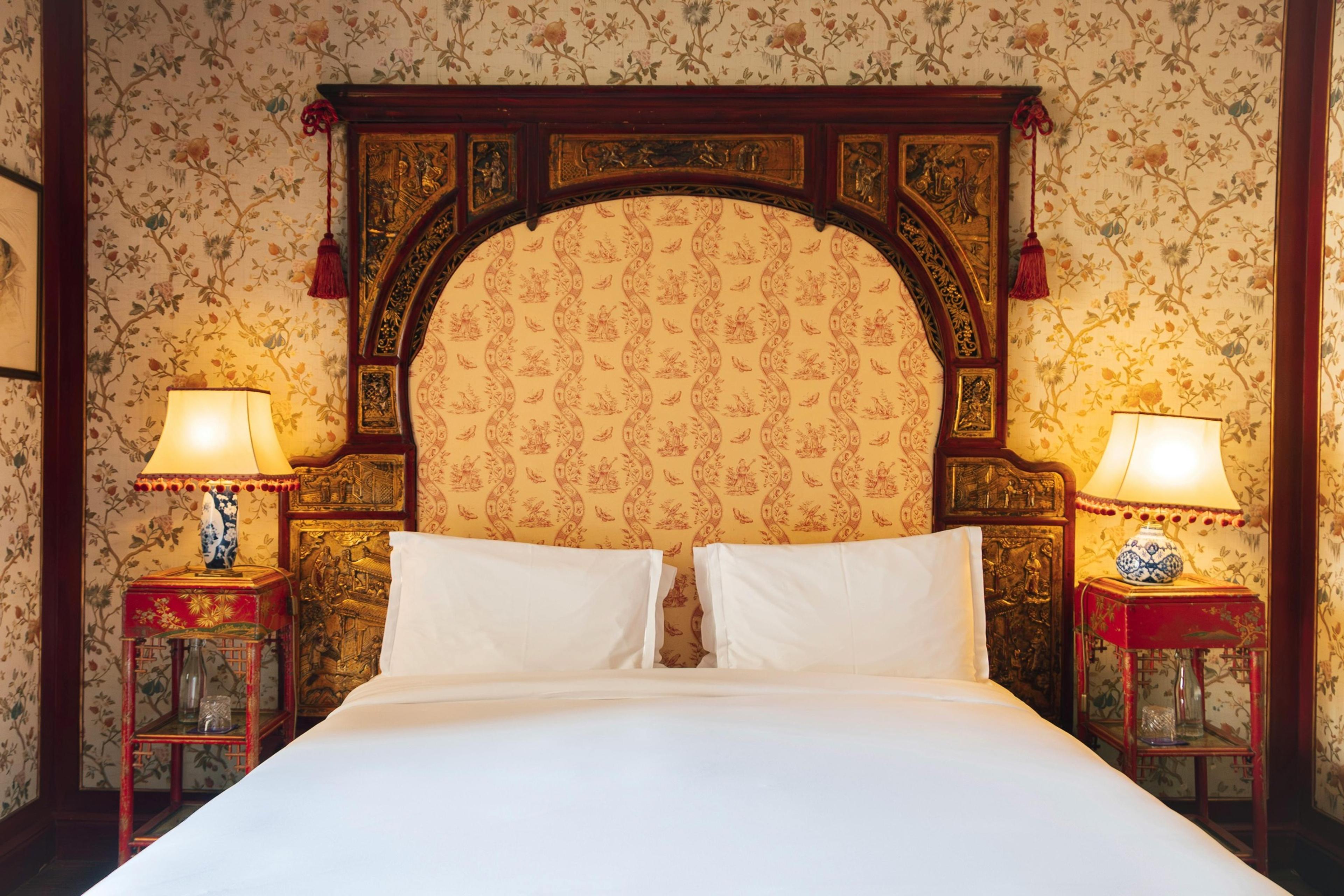 A richly carved and gilded antique bed with a scalloped headboard set against floral wallpaper, flanked by two matching lamps on bedside tables.