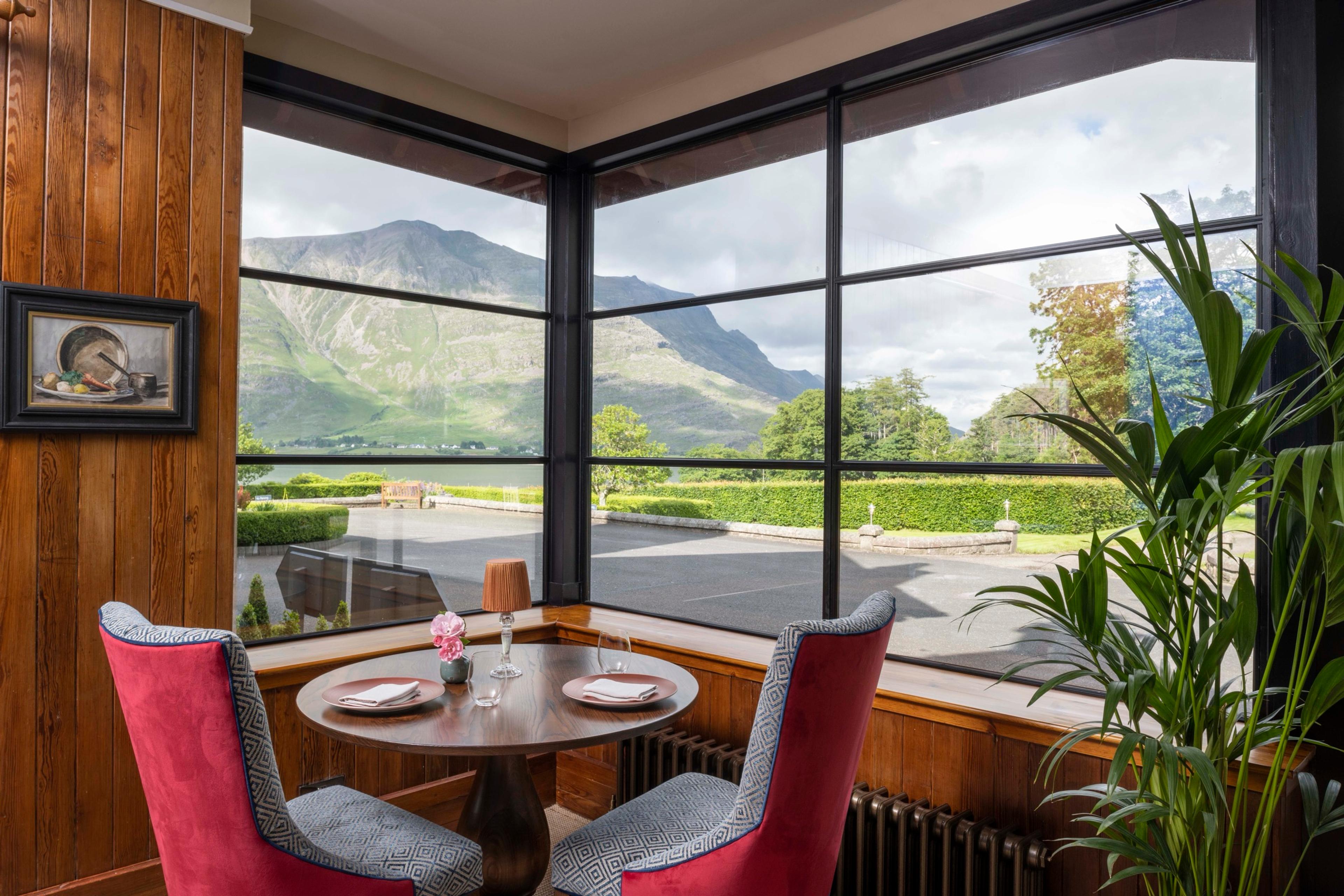 A corner dining table with two red chairs beside a wall of windows looking out over a lawn to mountains and a loch.