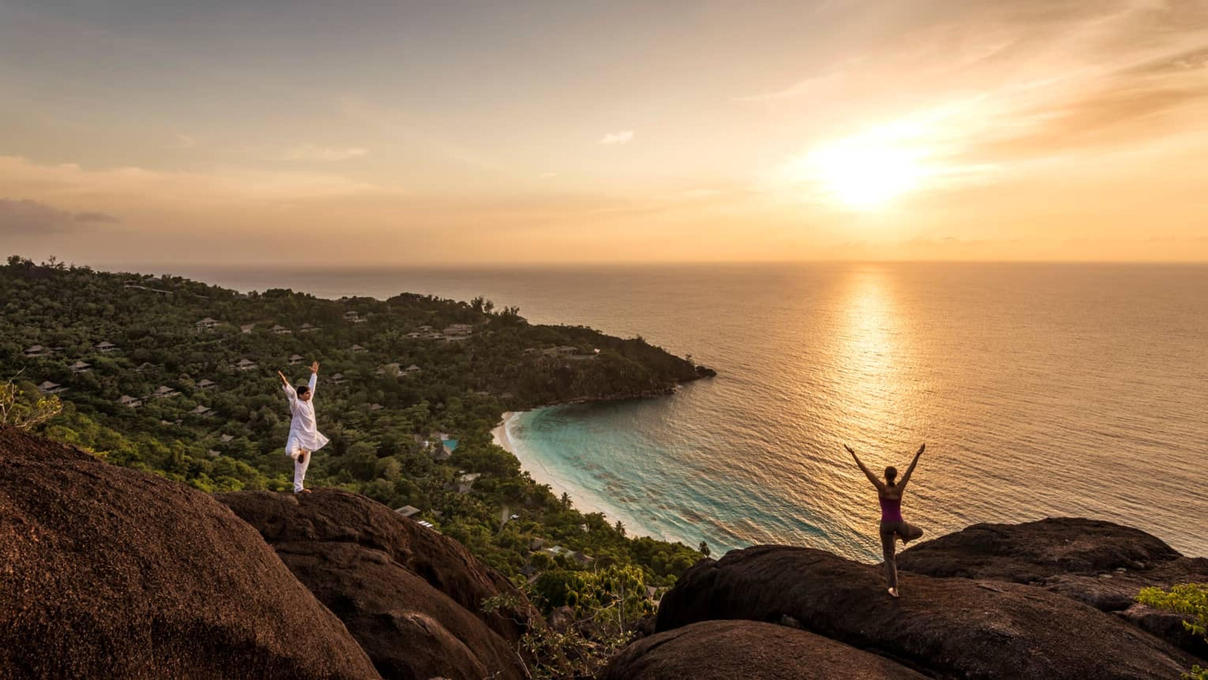 Two people silhouetted in a yoga pose on granite boulders above the coast at sunset.