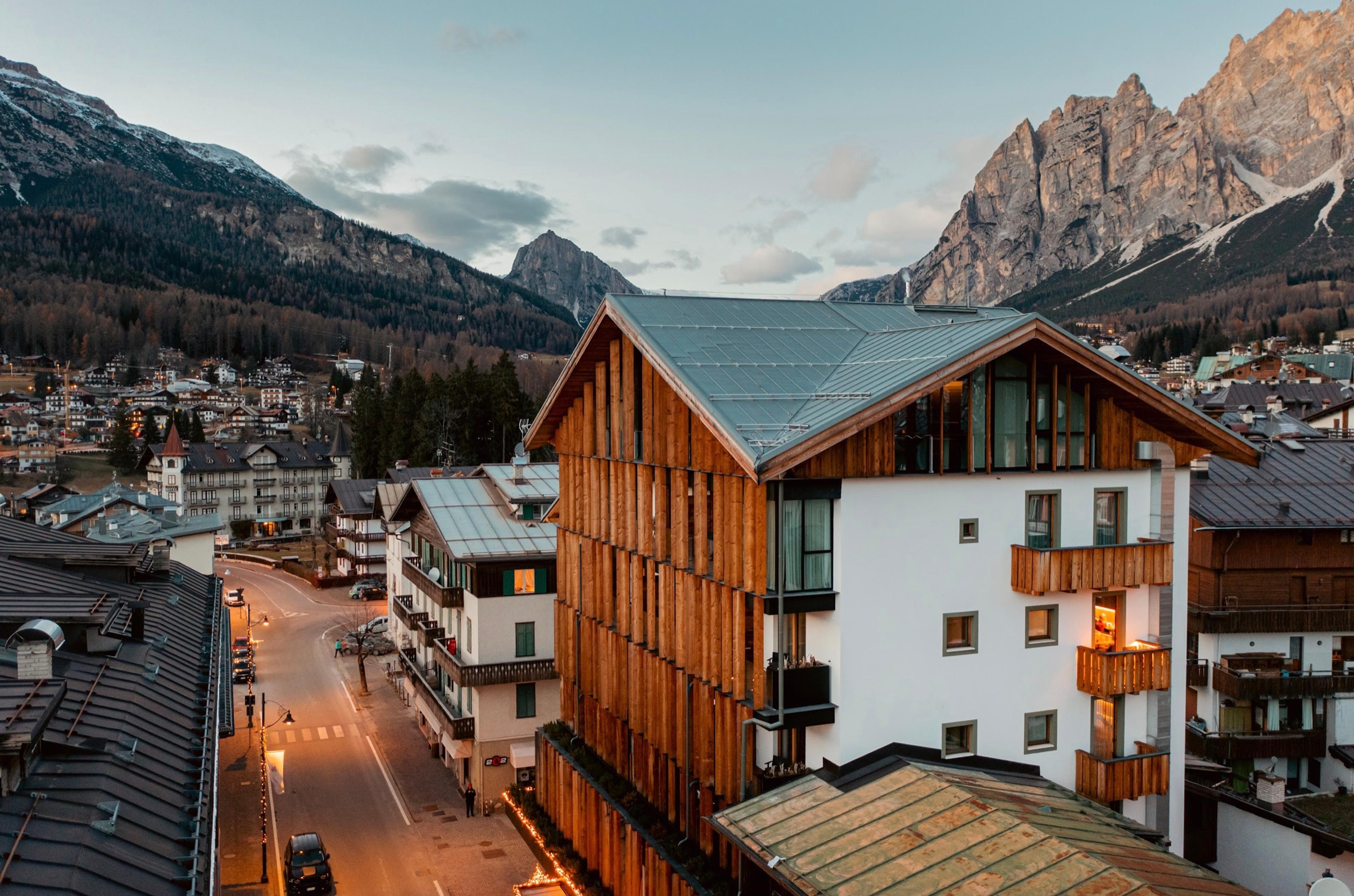 An aerial dusk view shows the hotel's timber and white facade lit against the mountains, above a street lined with lights.