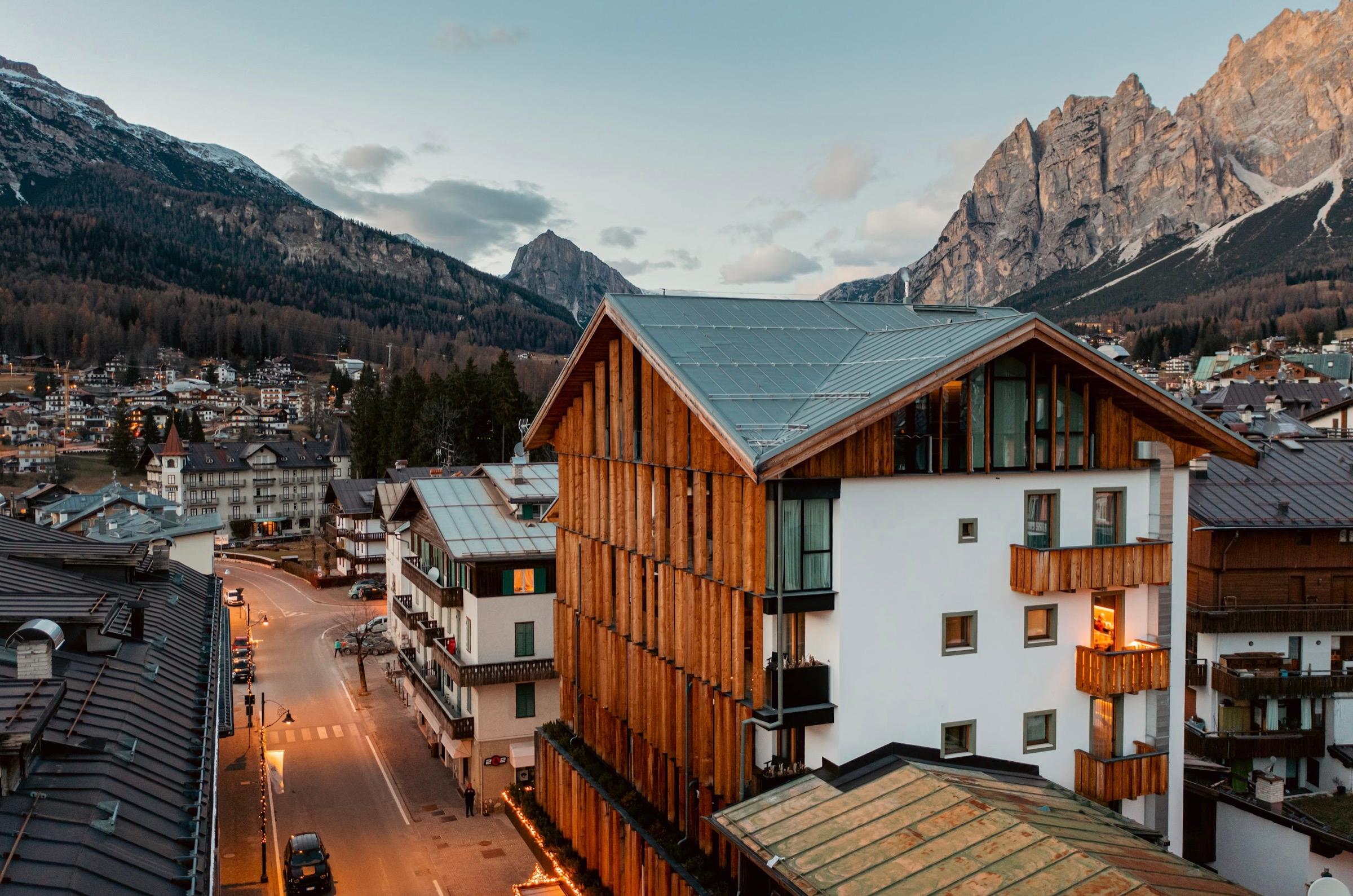 An aerial dusk view shows the hotel's timber and white facade lit against the mountains, above a street lined with lights.