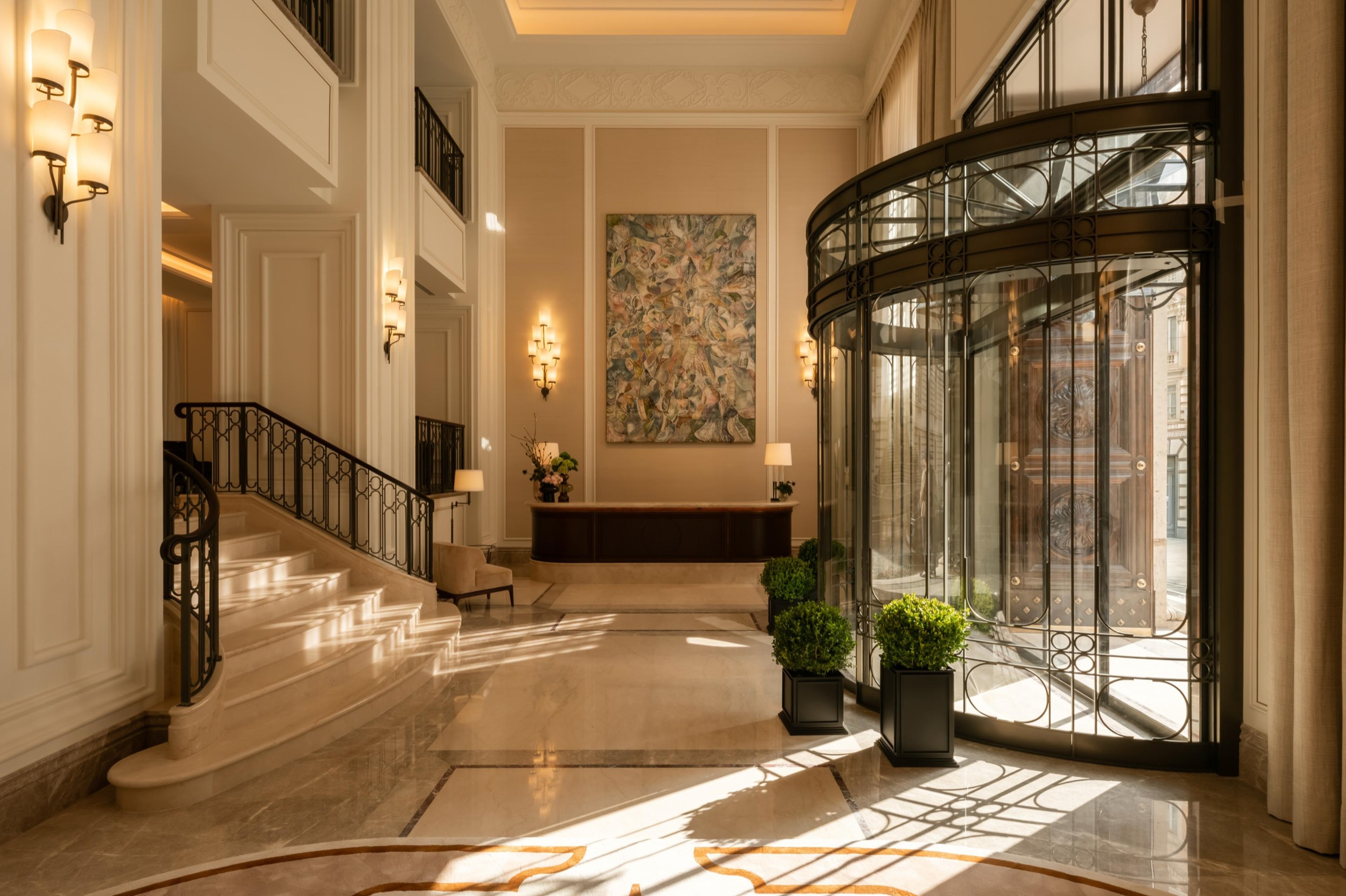 a hotel lobby with a curved marble staircase, a glass revolving door, potted topiary and a large abstract painting on the far wall