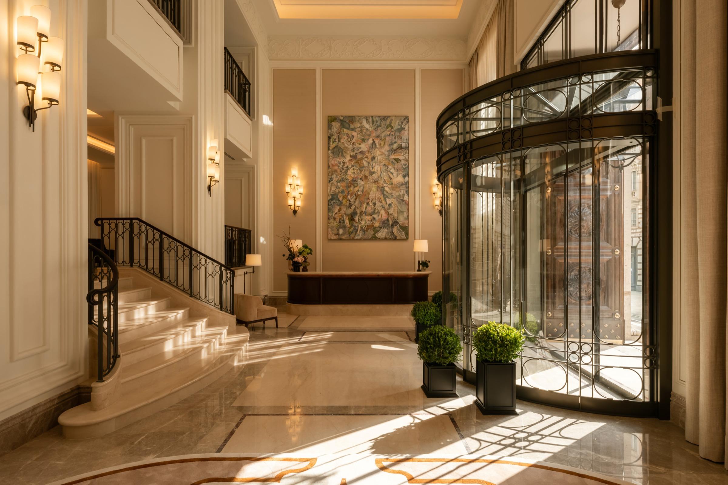 a hotel lobby with a curved marble staircase, a glass revolving door, potted topiary and a large abstract painting on the far wall
