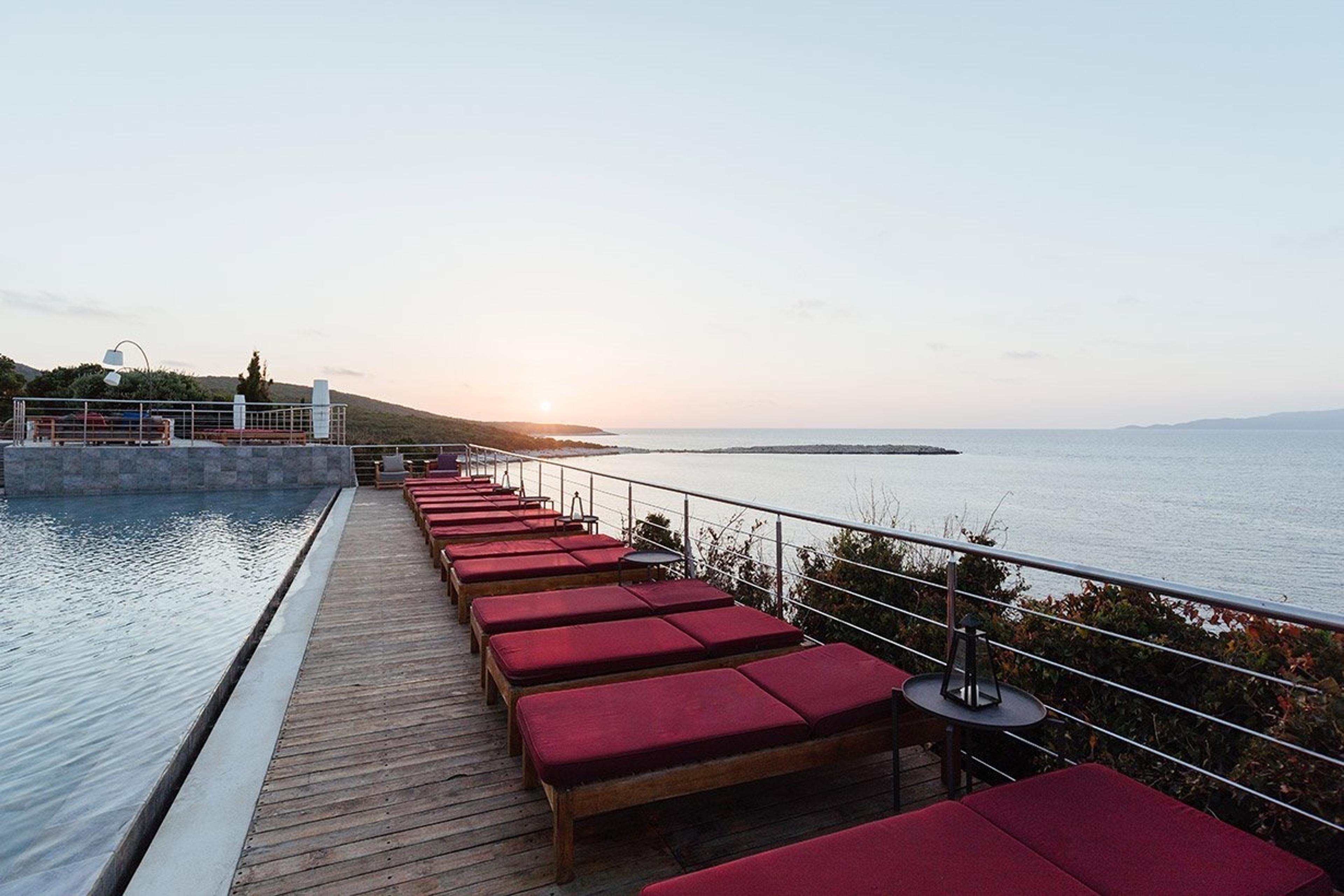 A row of red sun loungers along a railing overlooking the open sea at sunset.