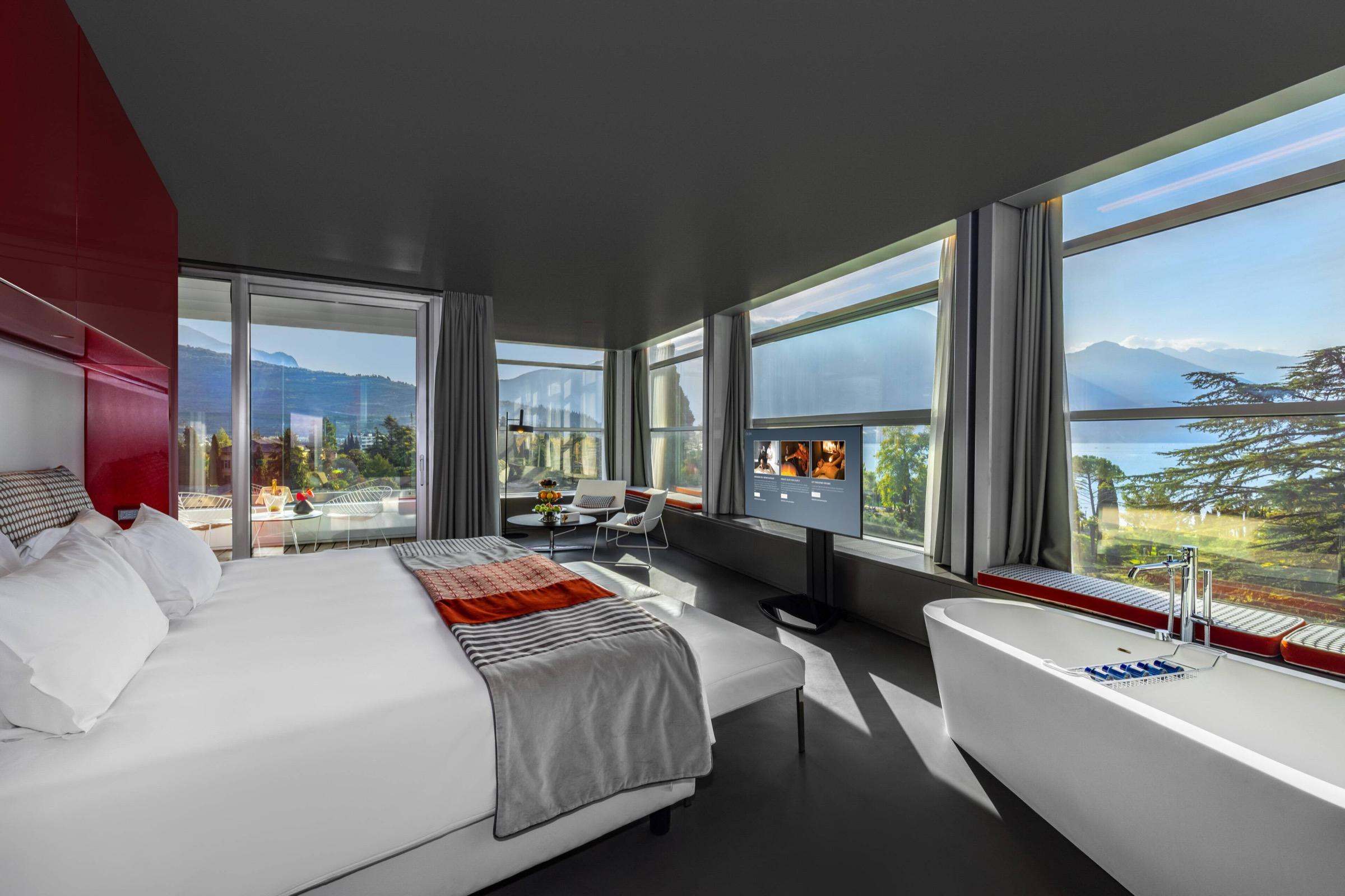 A hotel bedroom with a white bed and a freestanding bathtub set beside a wall of windows looking over trees, water and mountains.