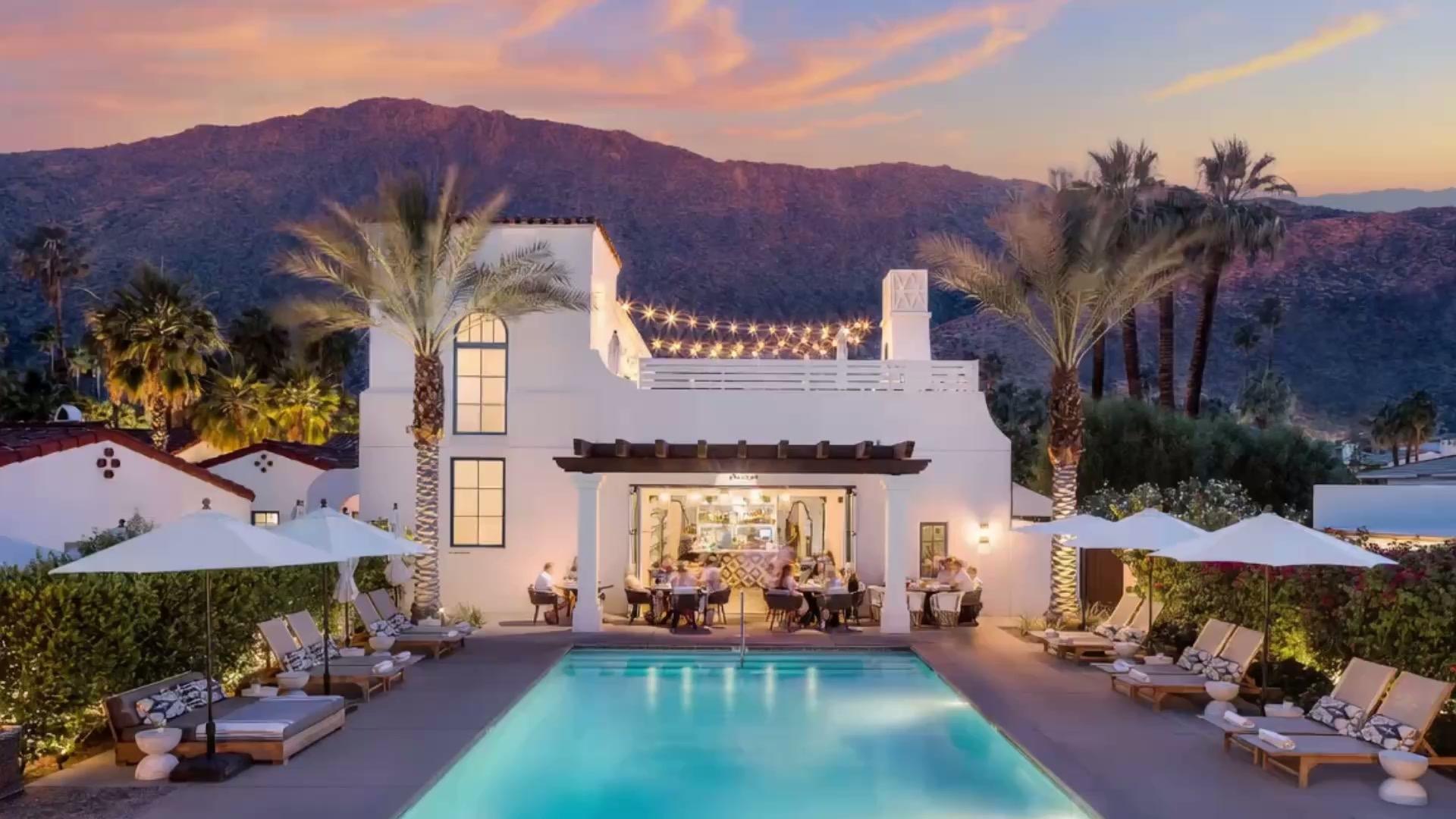 A white hotel building and swimming pool lit at sunset, with guests dining under a pergola bar and mountains behind.