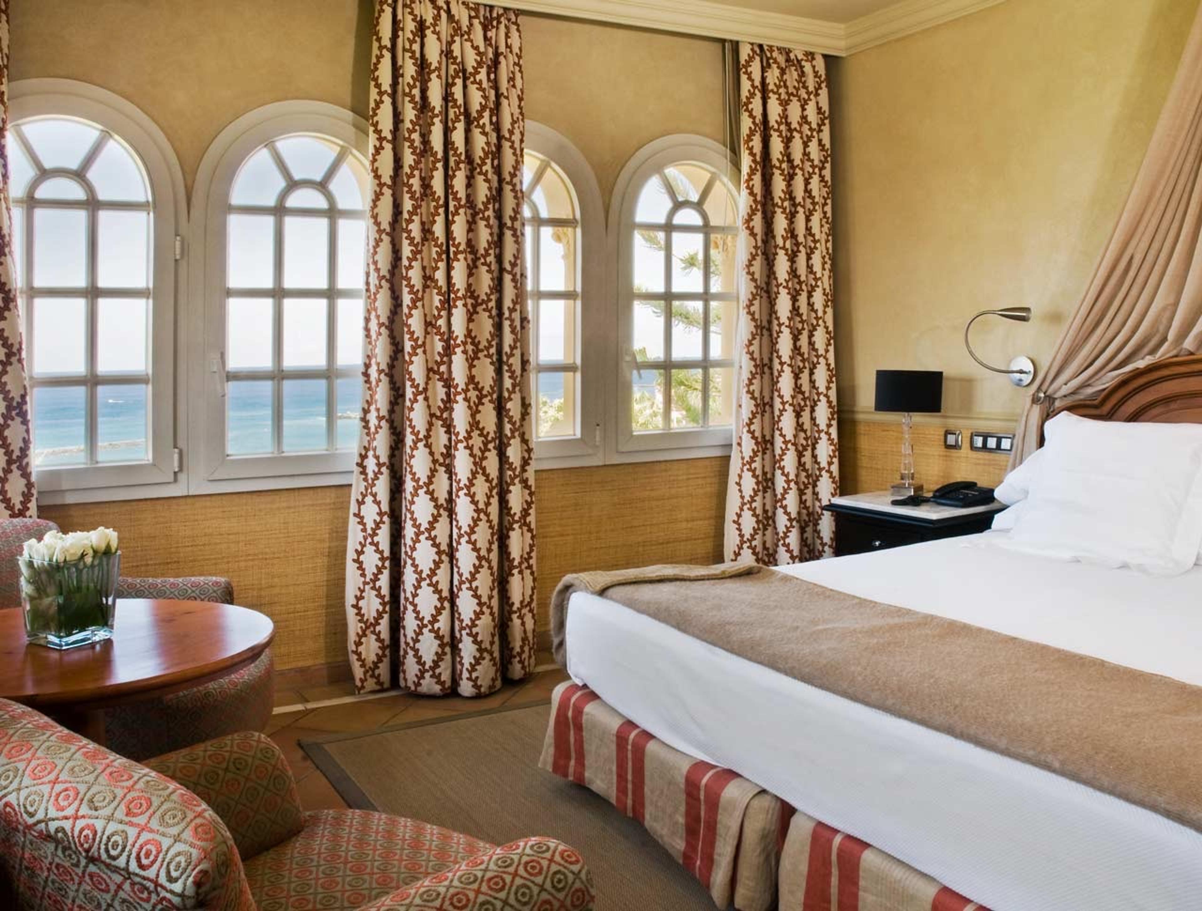 Three arched windows hung with patterned curtains looking out over the sea, beside a corner of a four-poster bed.