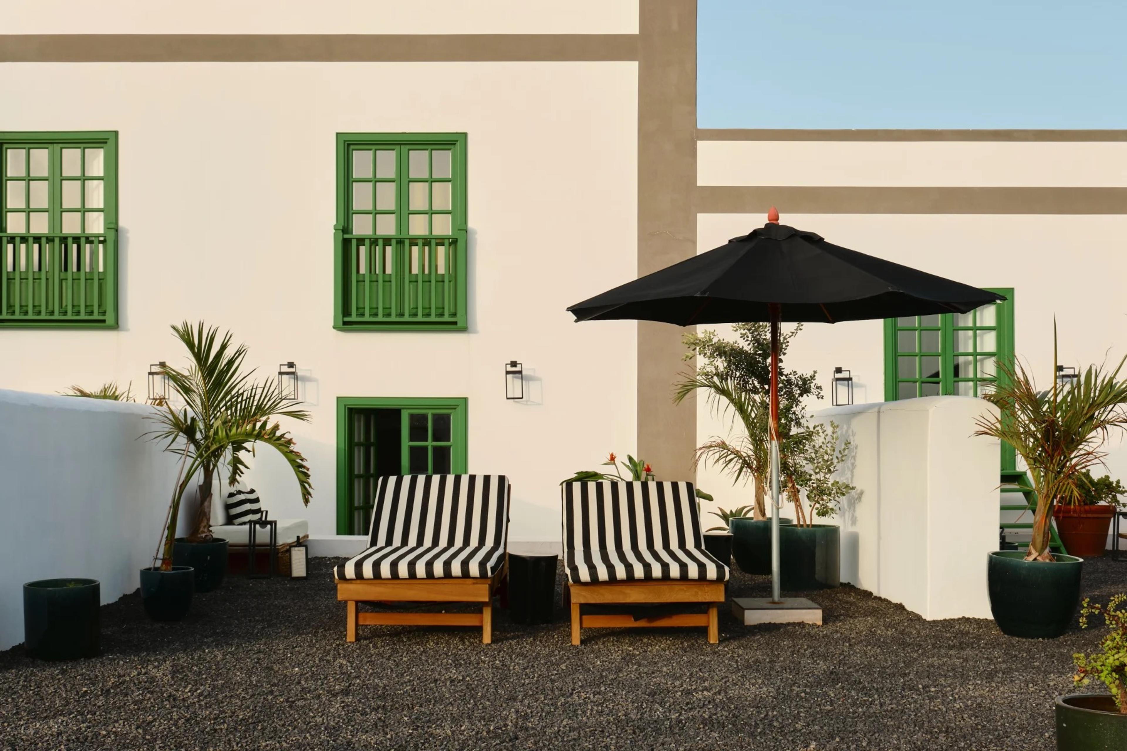 Two black and white striped sun loungers and a black umbrella on gravel in front of a white hotel facade with green-shuttered windows and doors.