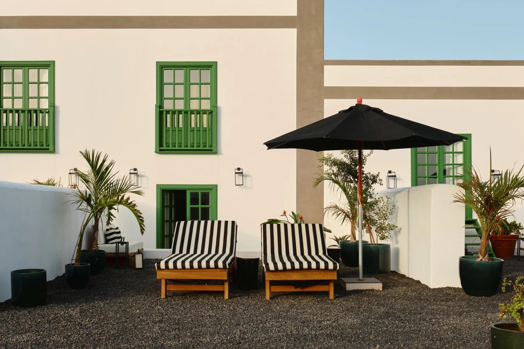Two black and white striped sun loungers and a black umbrella on gravel in front of a white hotel facade with green-shuttered windows and doors.