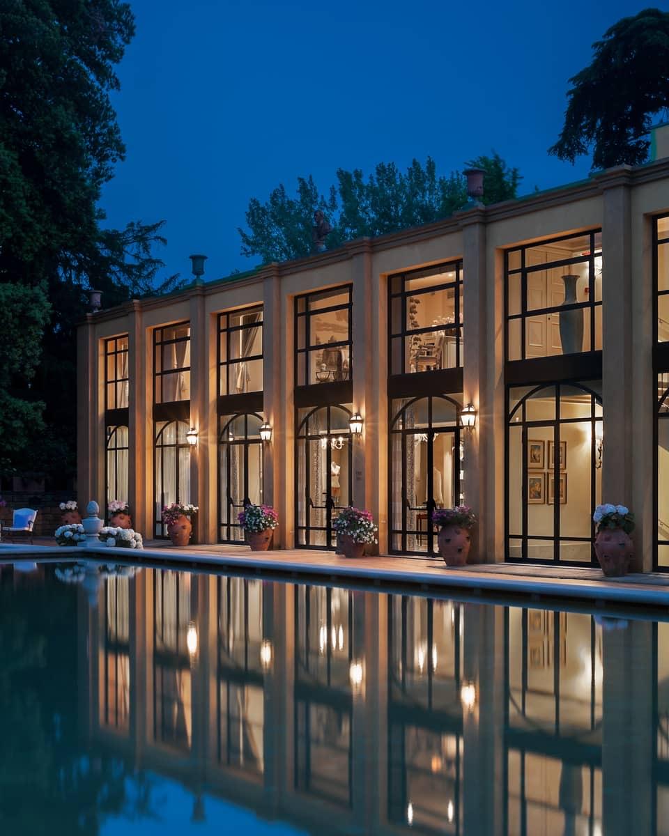 A two-storey building with a row of tall arched windows lit from inside, reflected in a pool at dusk.