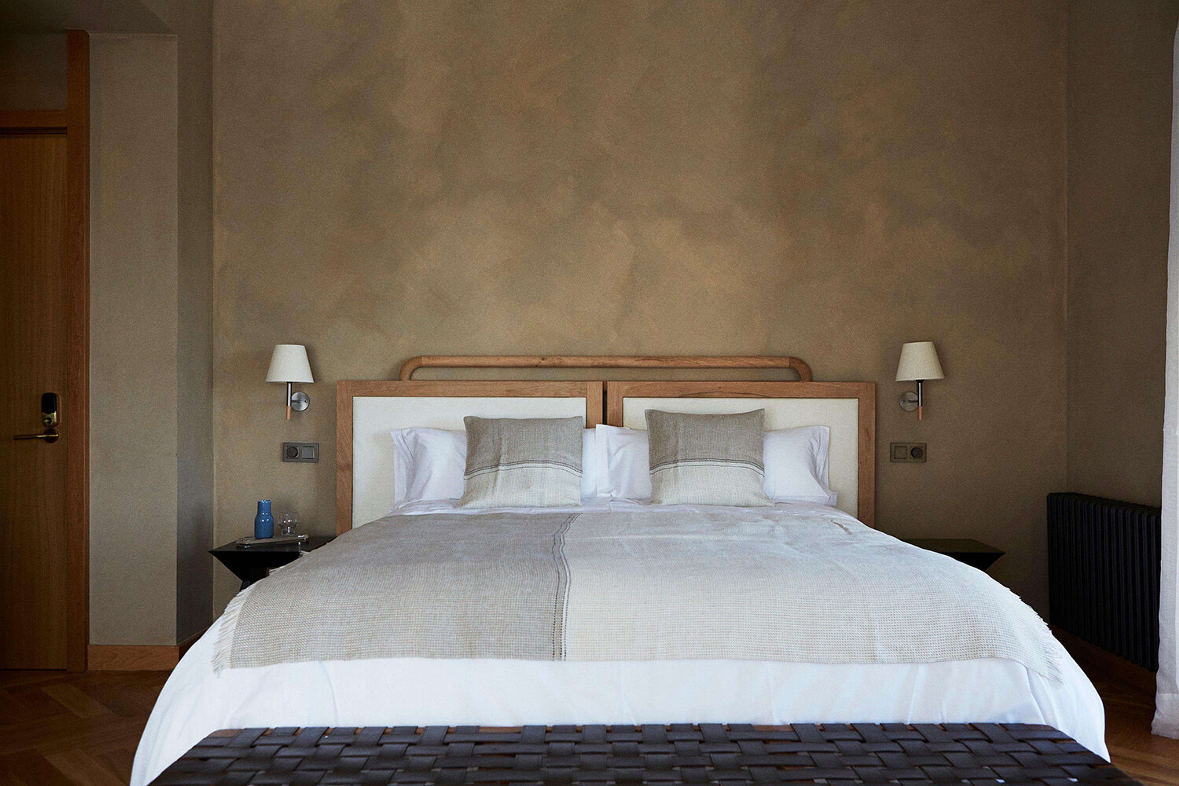 a neatly made double bed with grey cushions between two wall lamps, a wooden door and a radiator to the sides