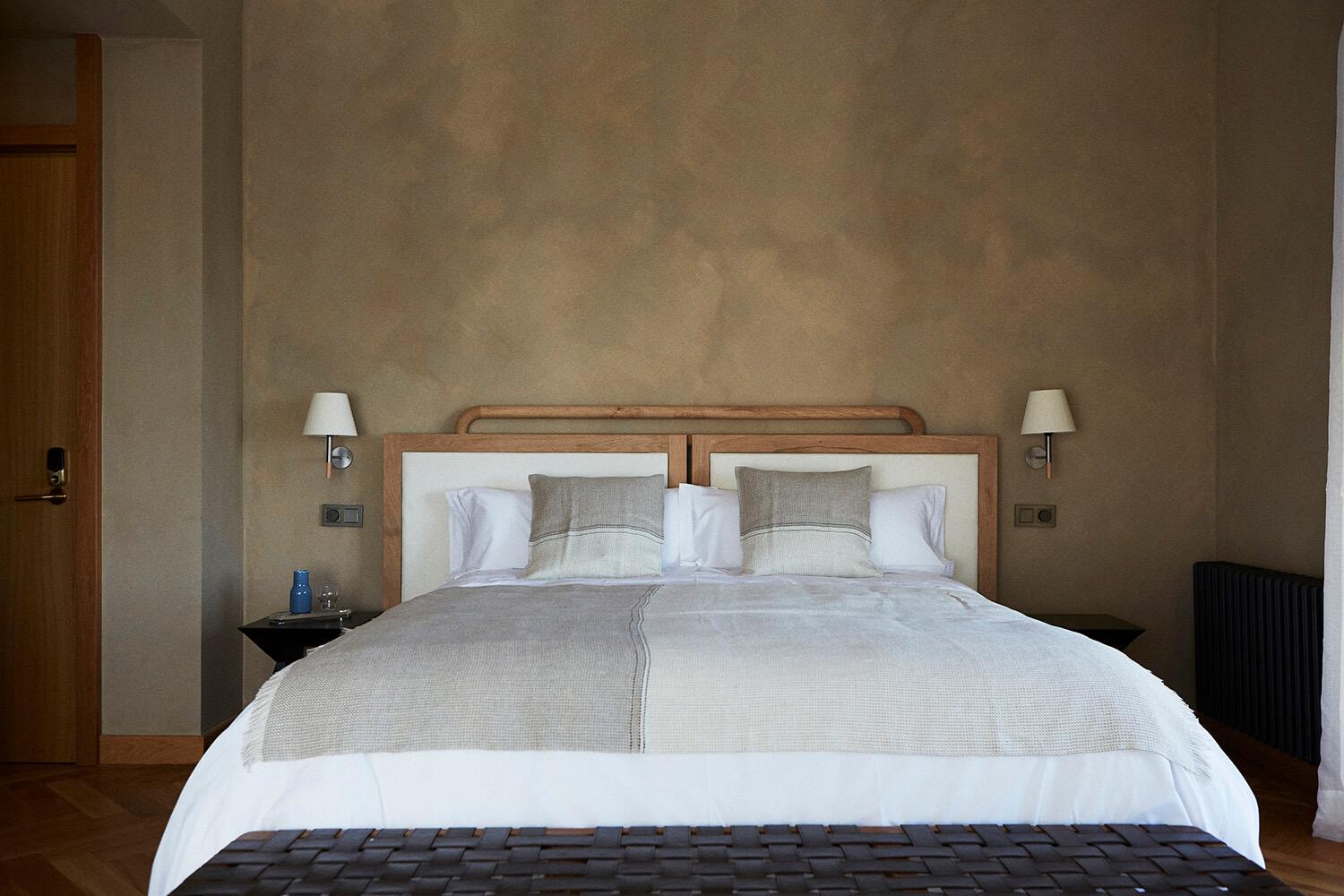 a neatly made double bed with grey cushions between two wall lamps, a wooden door and a radiator to the sides