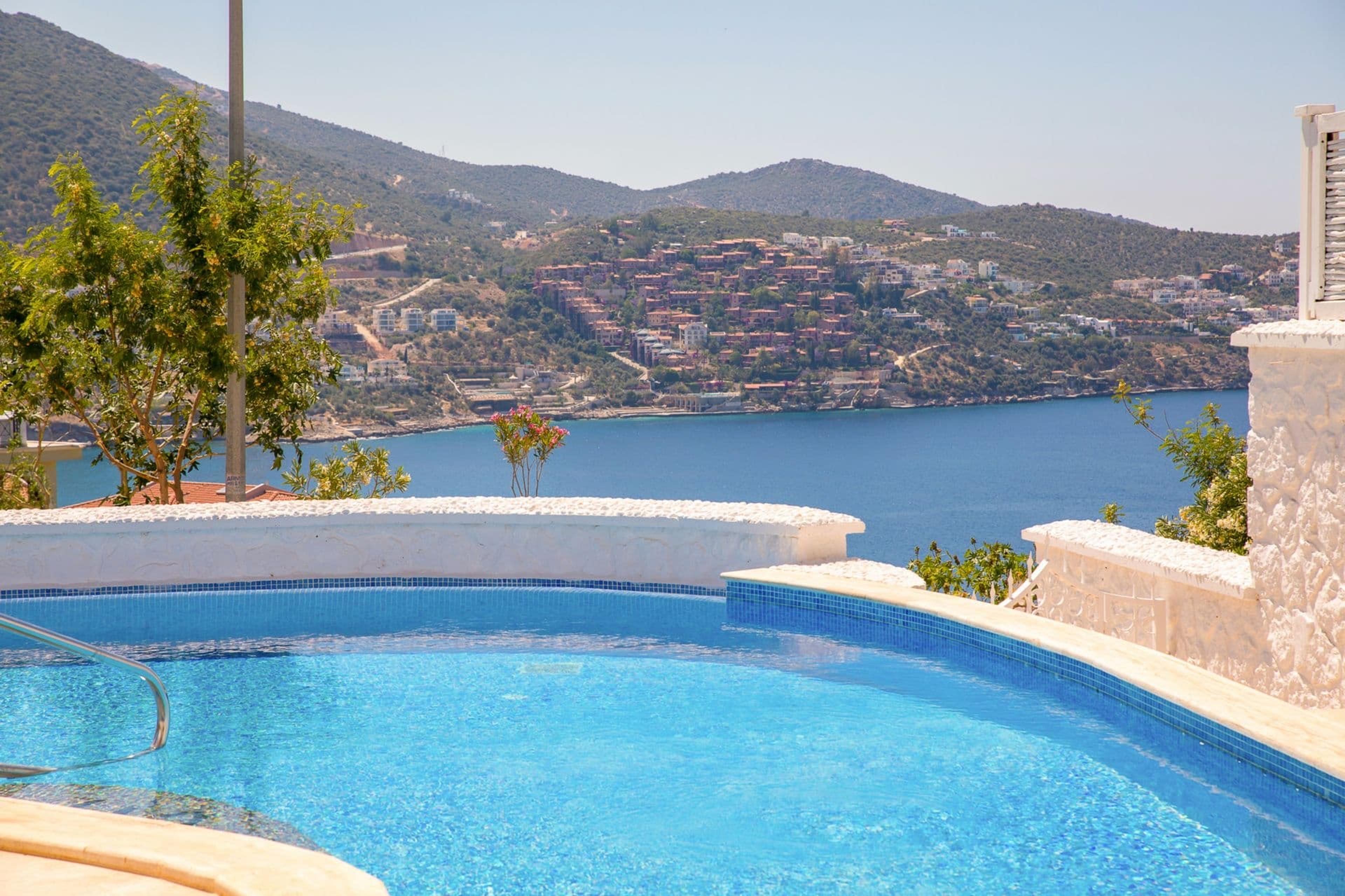 The curved edge of a swimming pool looks out over a bay to hills and a hillside town beyond.