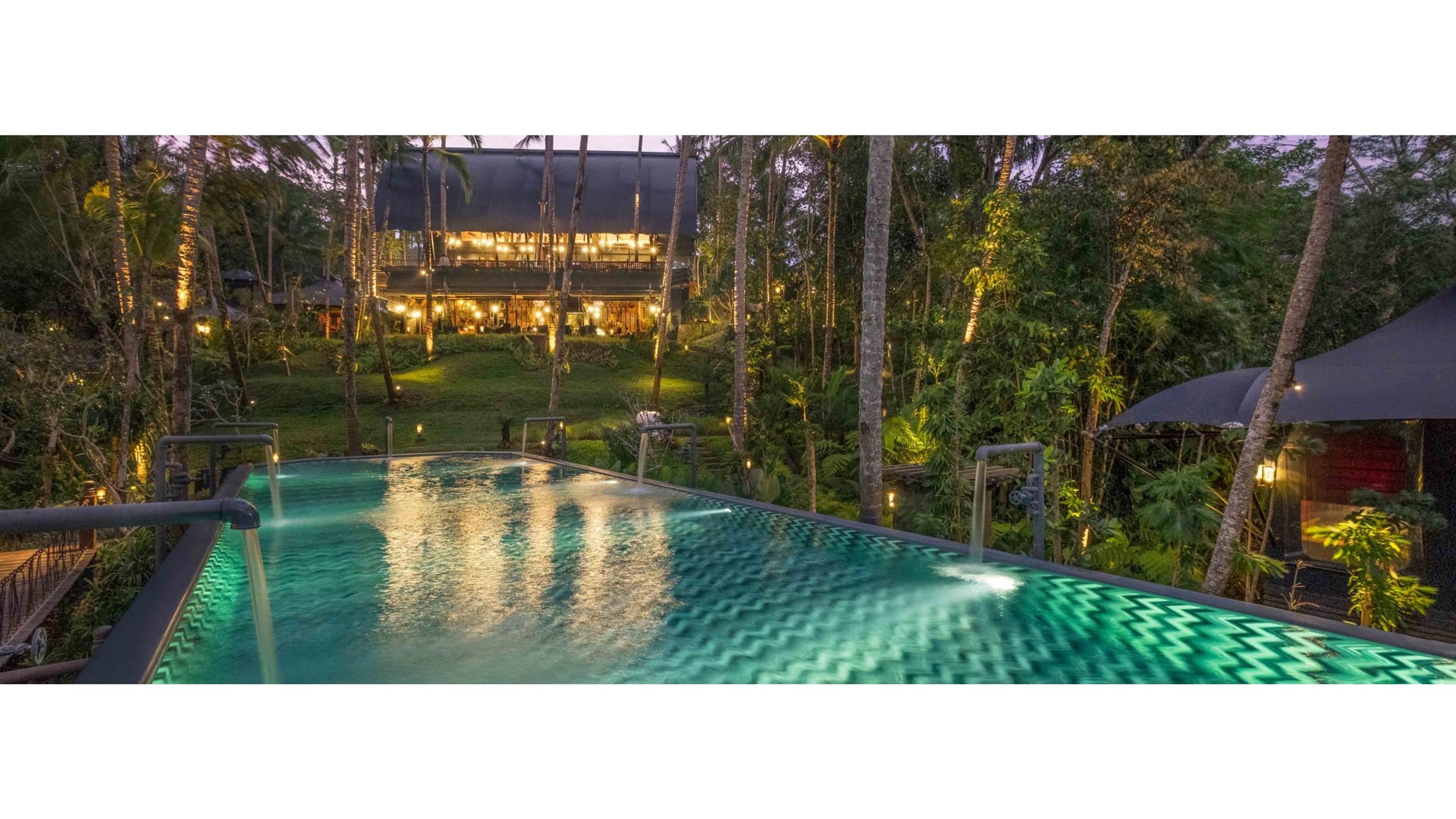 An infinity pool with illuminated water jets in the foreground, and a two-storey open-sided building lit from within beyond it, surrounded by palm trees at dusk.