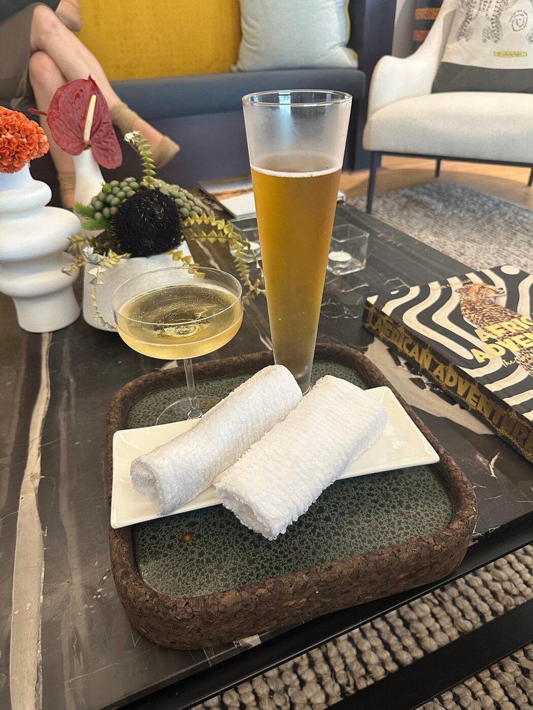 Two rolled white hand towels on a white plate on a cork tray, beside a coupe of sparkling wine and a tall glass of beer on a dark marble table.