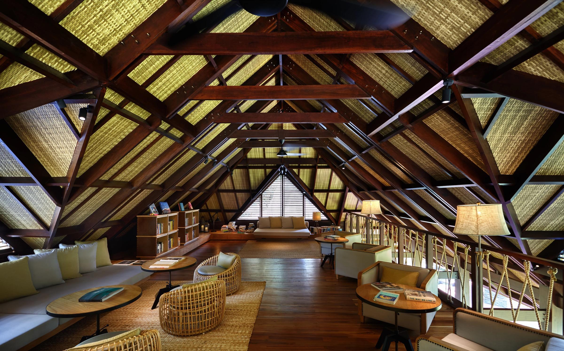 A loft room with a steep thatched roof, exposed wooden beams, woven armchairs, a sofa and shelves of books.