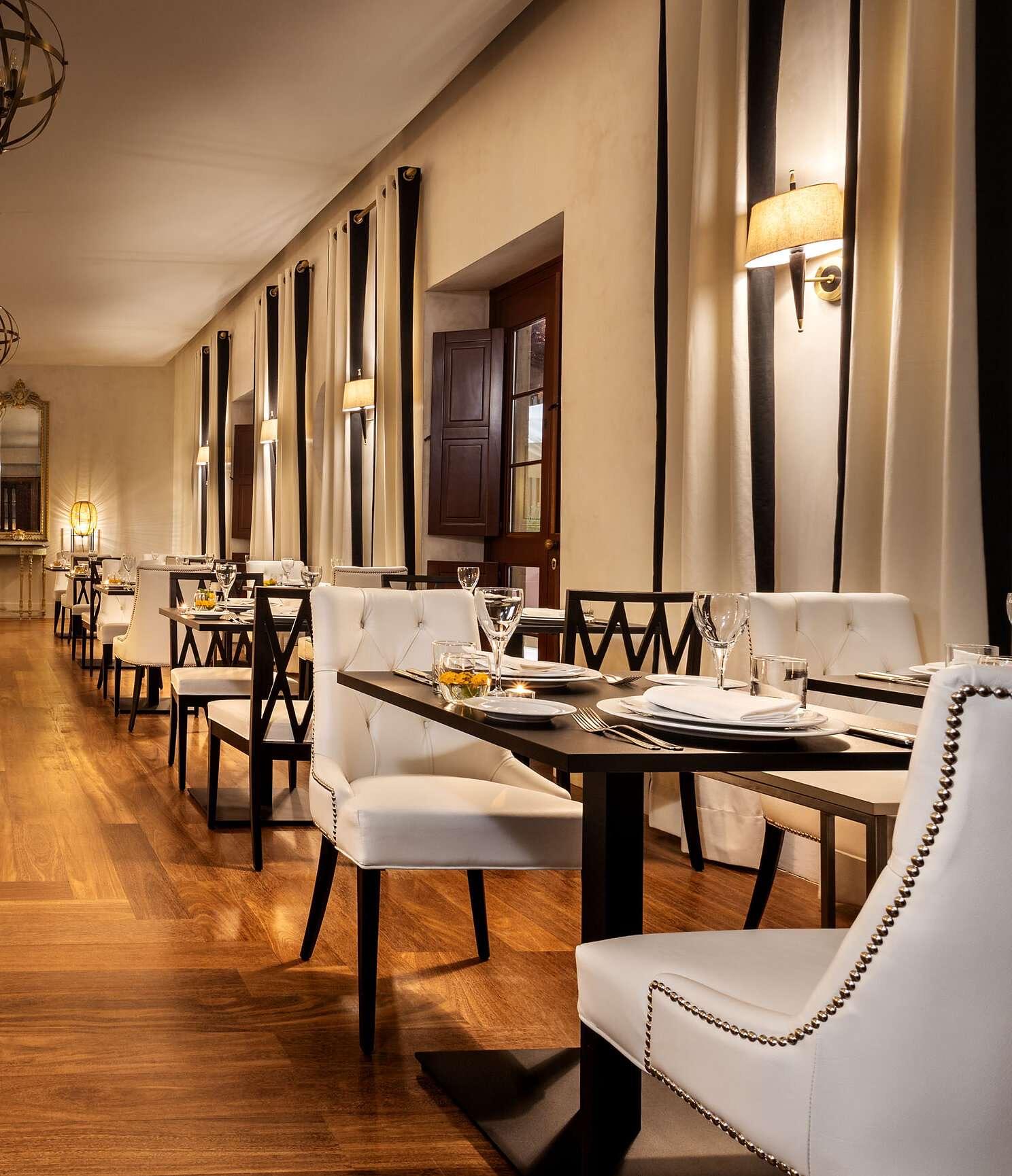 A dining room with a long wooden floor, tables laid with white plates and glasses, and white upholstered chairs under curtains with black stripes.
