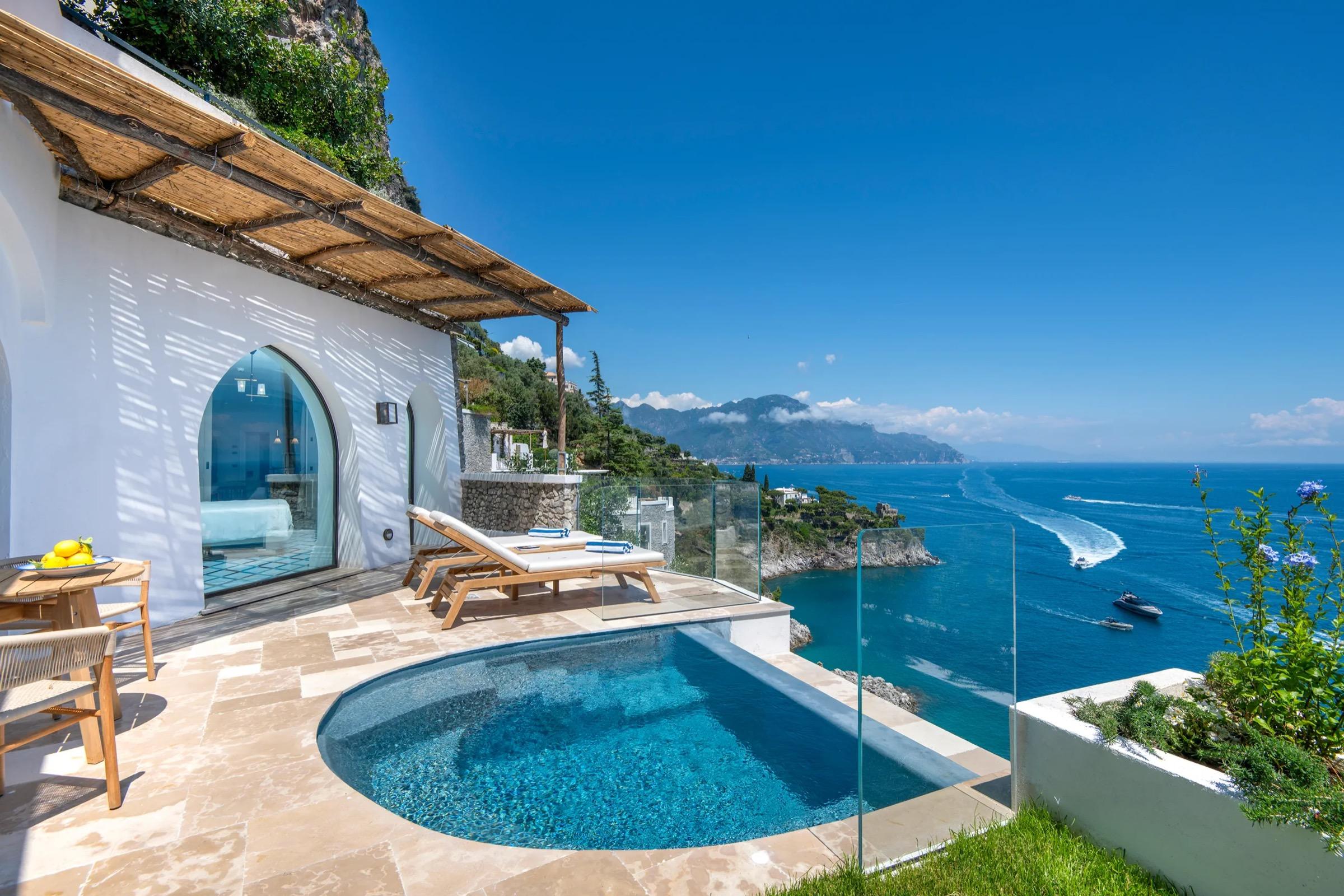 A round plunge pool on a stone terrace beside a whitewashed building, with two sun loungers, a dining table and a glass balustrade overlooking the sea.