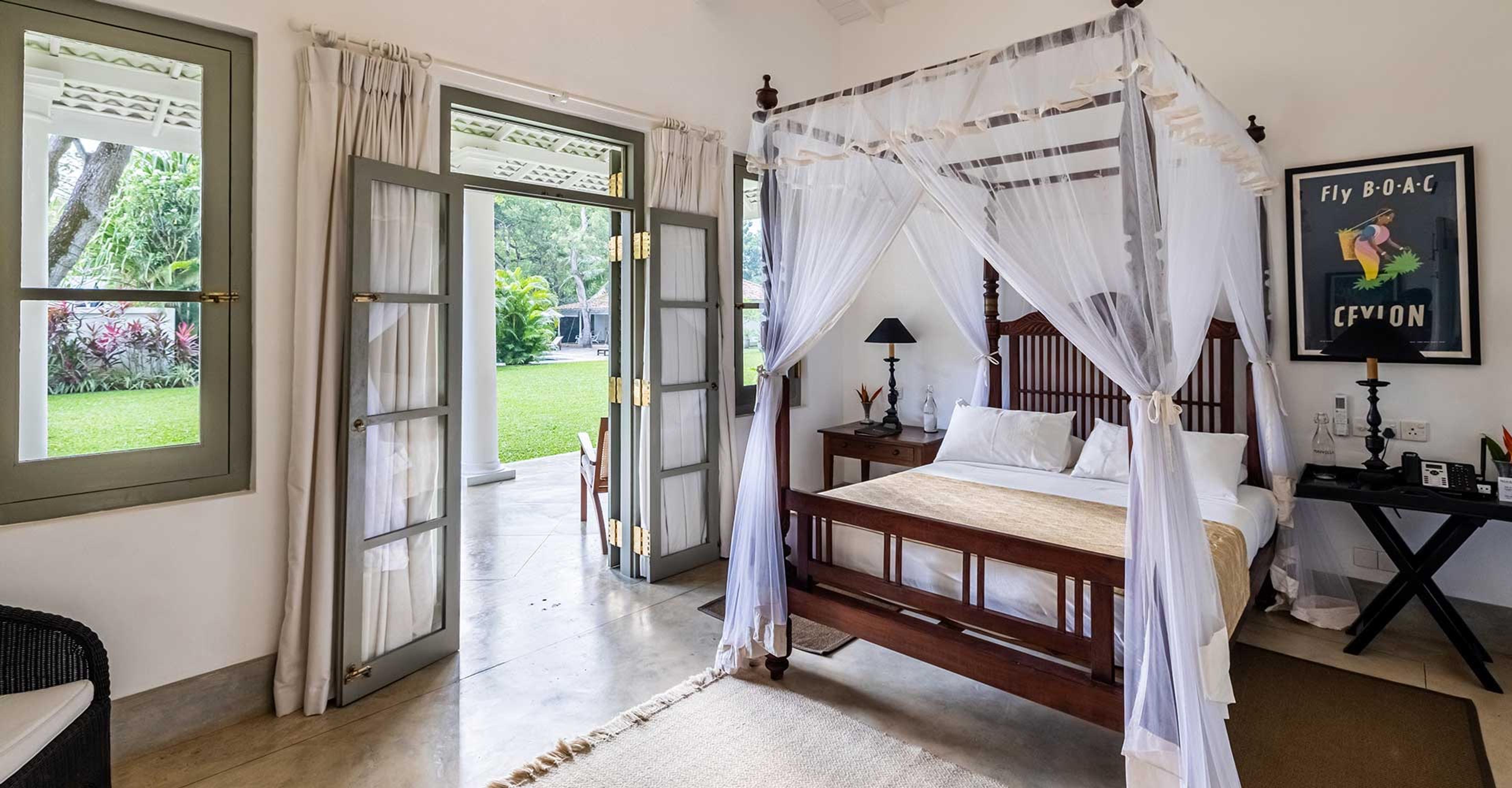 A bedroom with a canopy bed under white netting, a framed Fly BOAC Ceylon poster on the wall, and folding doors open onto a lawn.