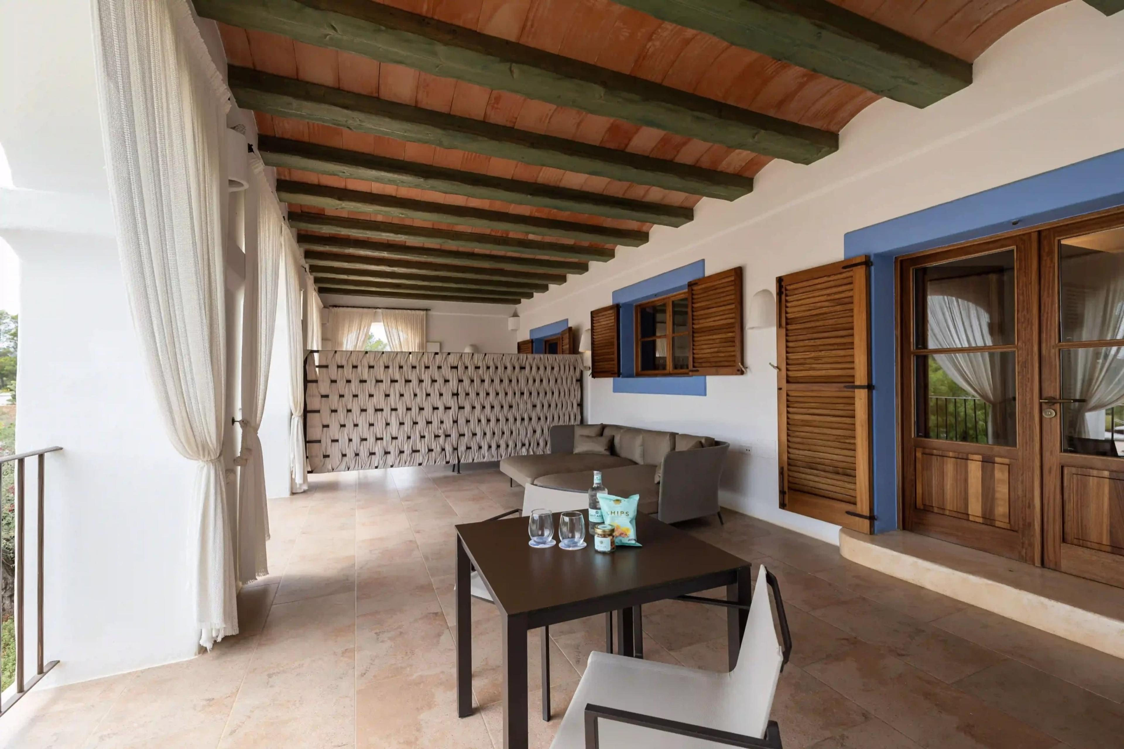 A covered terrace running the length of the building, its terracotta-beamed ceiling and blue-trimmed shutters and doors leading off it, with a sofa and a table set with drinks.