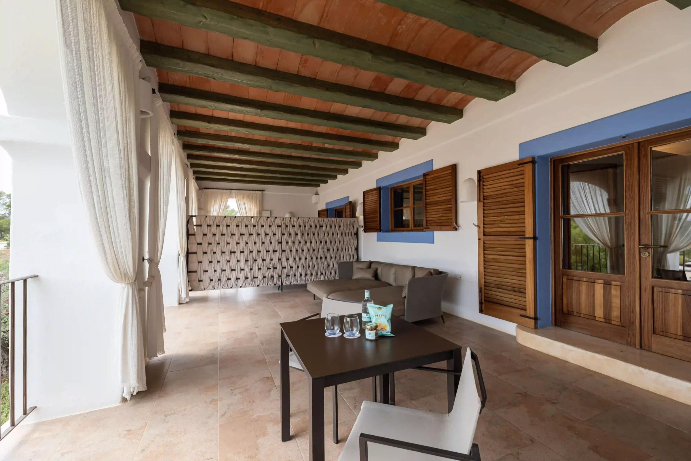 A covered terrace running the length of the building, its terracotta-beamed ceiling and blue-trimmed shutters and doors leading off it, with a sofa and a table set with drinks.