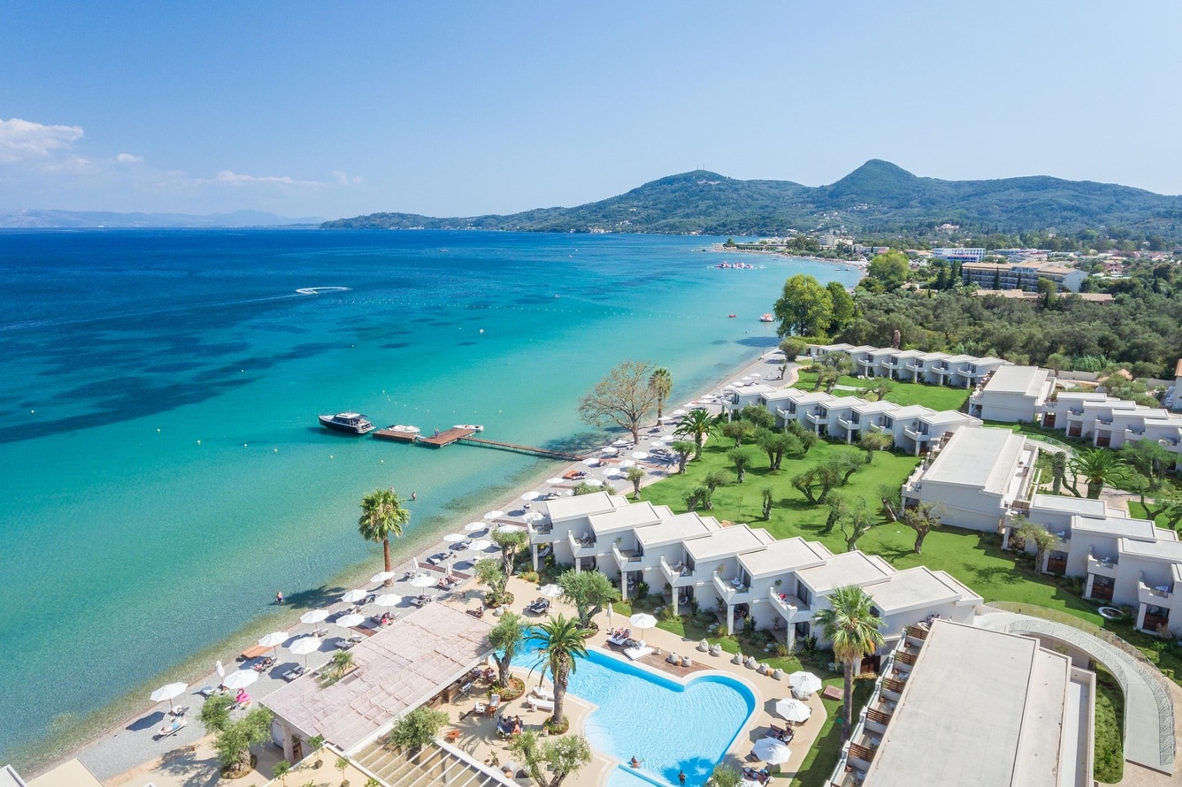 An aerial view of a seafront resort with a heart-shaped pool, sunbeds along a pebble beach, and rows of low-rise villas set among lawns and olive trees, with hills and a bay beyond.
