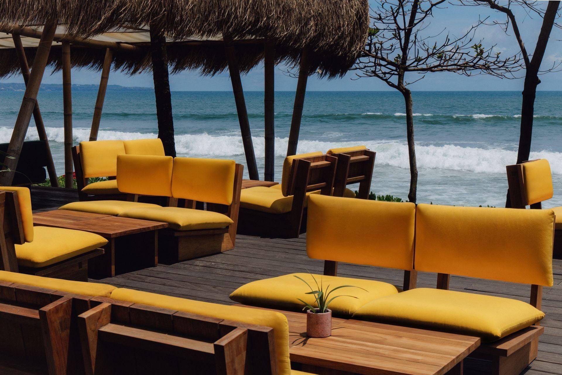 Empty yellow cushioned benches face the ocean under a thatched roof, a bare tree and waves breaking beyond.