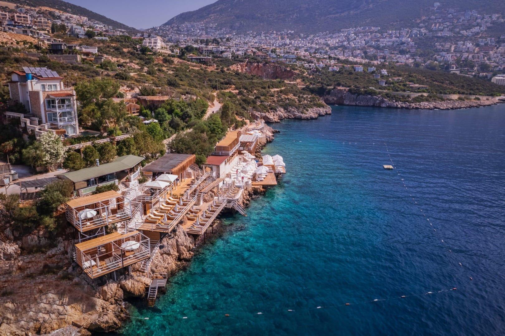 An aerial view of the hotel's cliffside sundeck and cabanas above the sea, with the town of Kalkan spread across the hills behind.