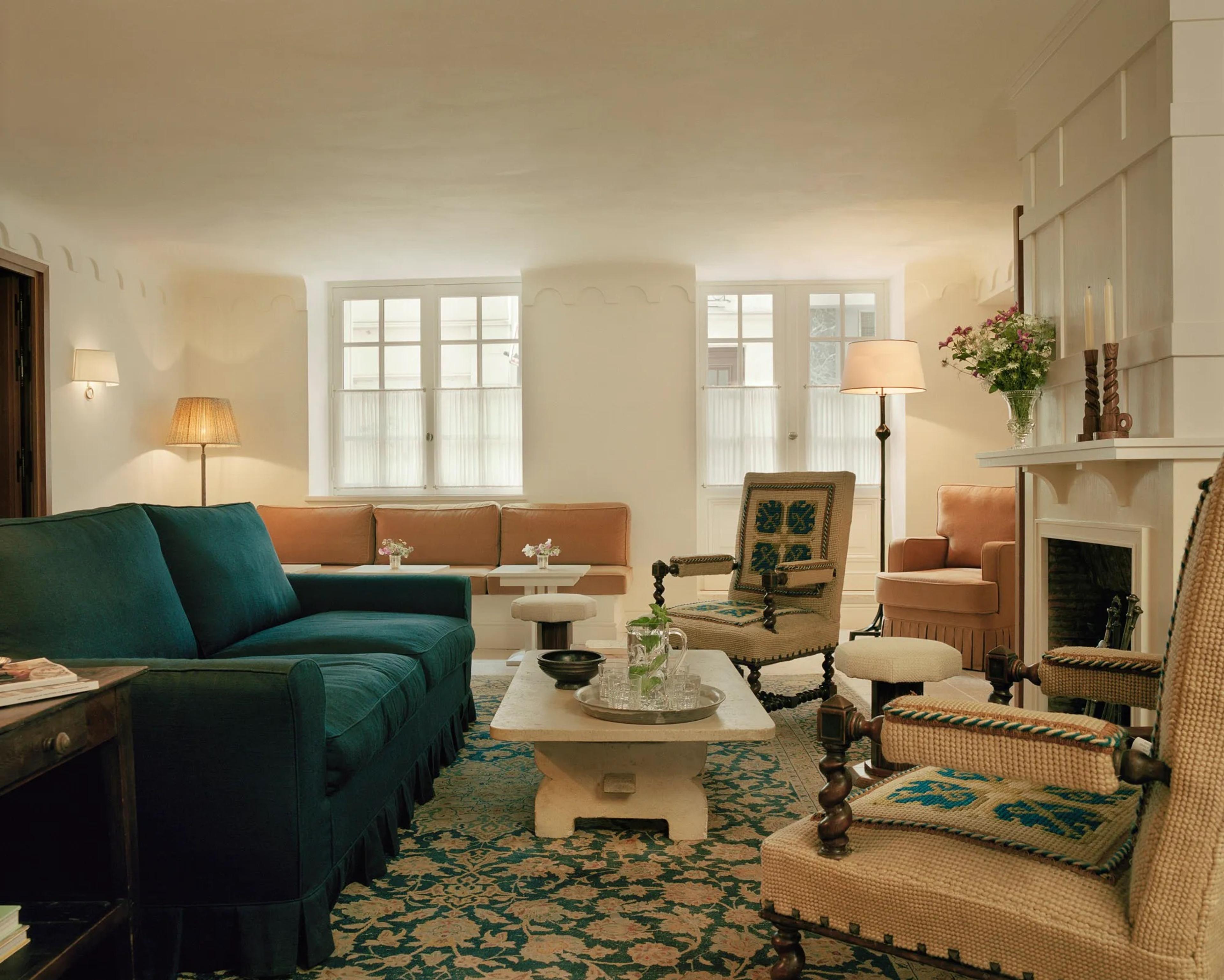 A salon with green velvet sofas, a fireplace and tall windows, books and flowers on a low table.