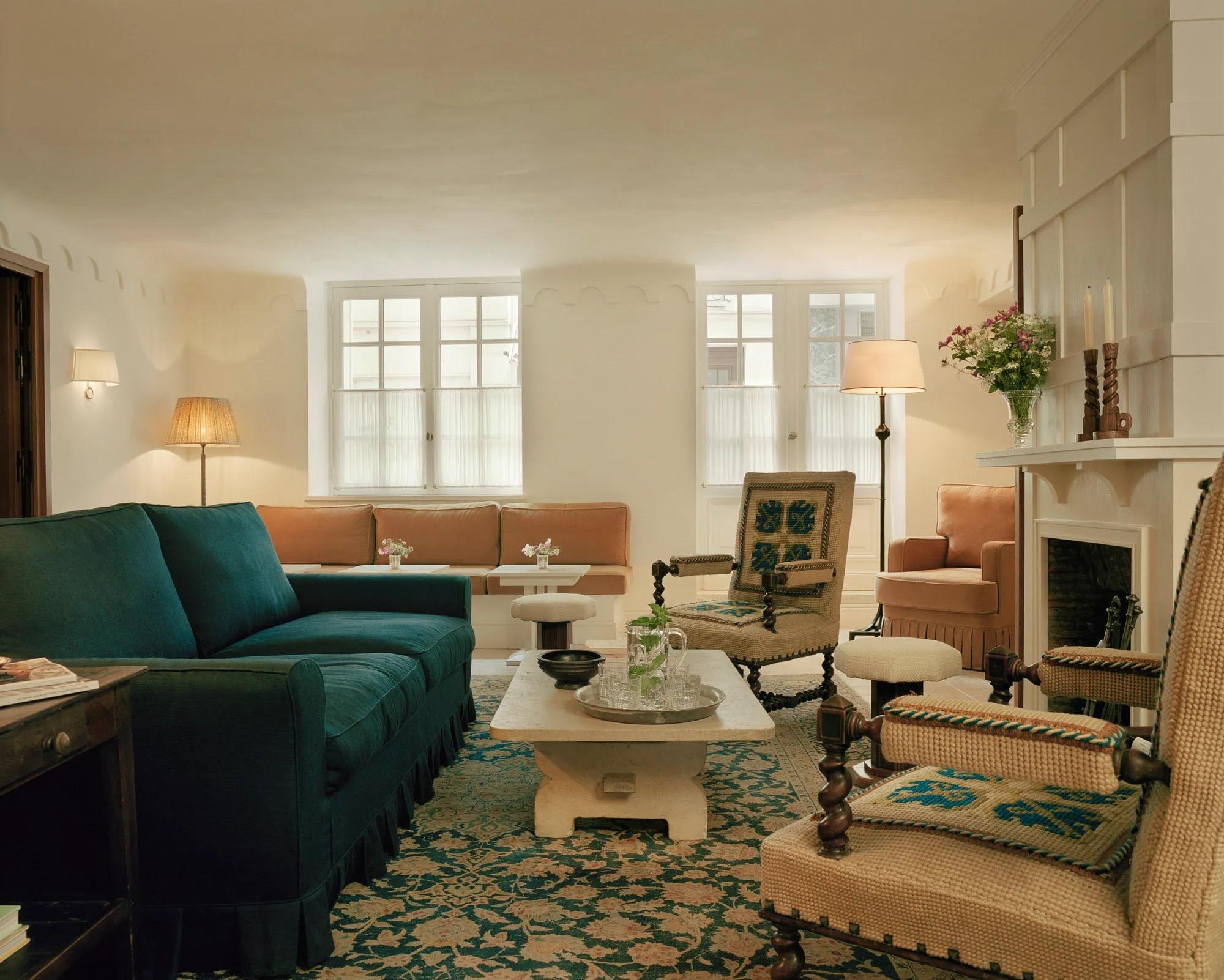A salon with green velvet sofas, a fireplace and tall windows, books and flowers on a low table.