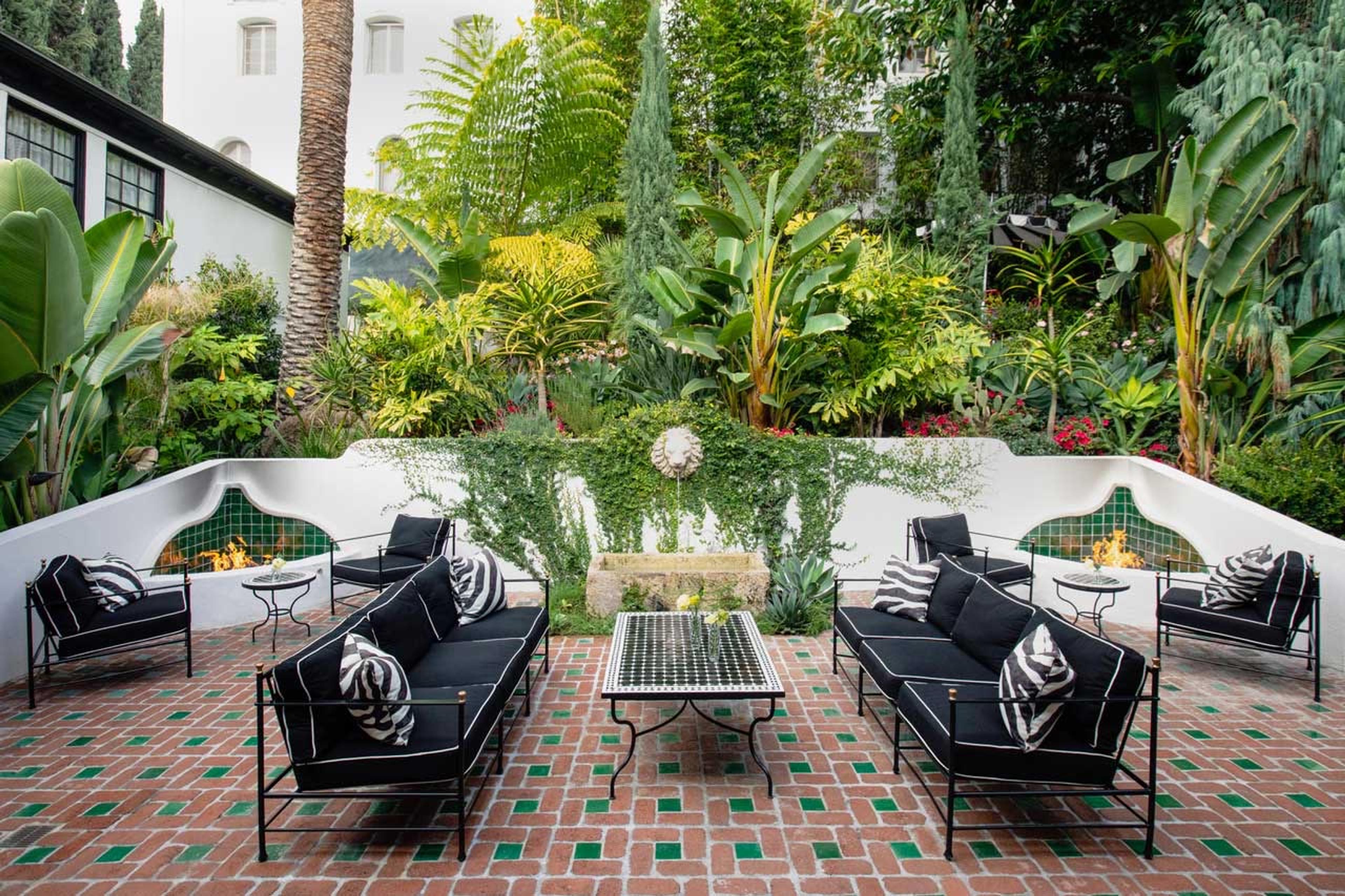 A courtyard with black and white striped sofas and armchairs around a mosaic coffee table, two built-in fireplaces, and dense tropical planting against a white building with arched windows.