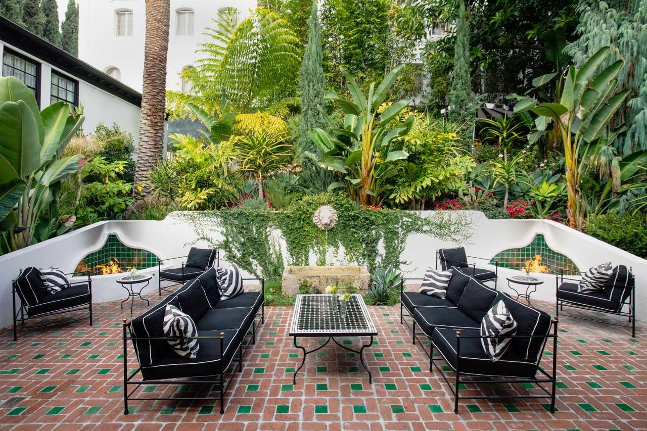 A courtyard with black and white striped sofas and armchairs around a mosaic coffee table, two built-in fireplaces, and dense tropical planting against a white building with arched windows.