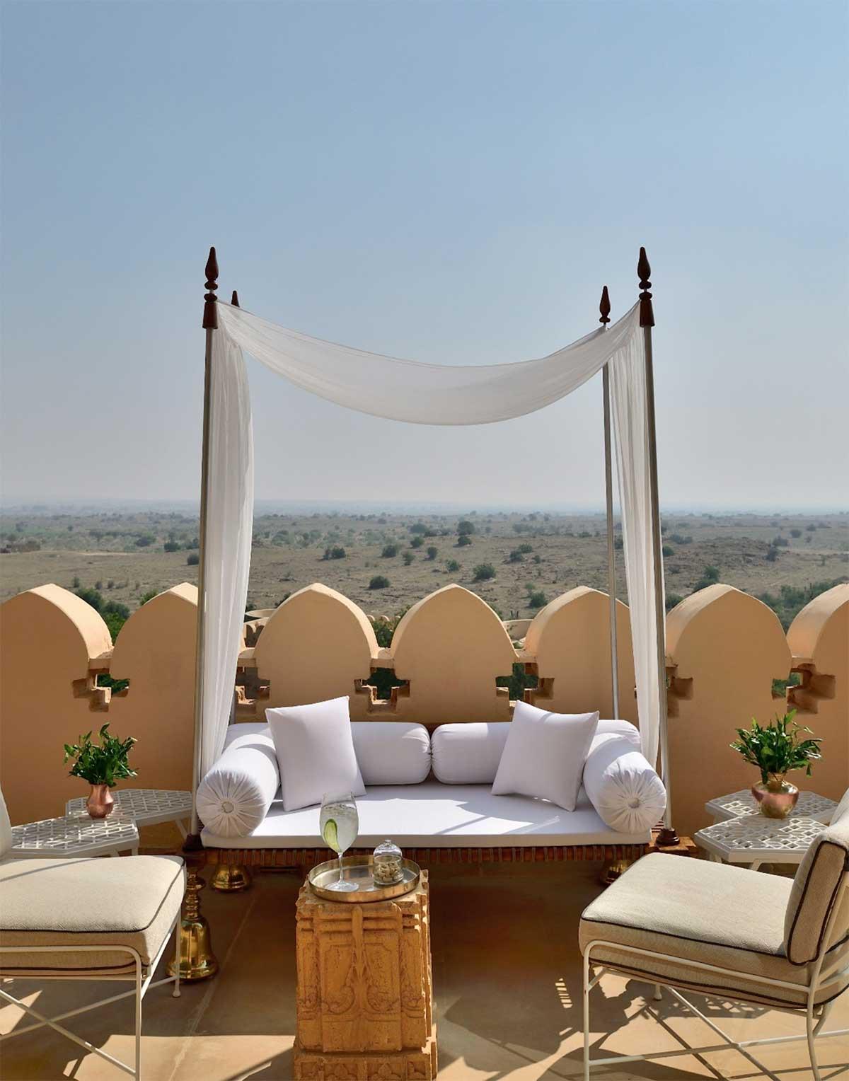 a white canopied daybed on a rooftop terrace, with cushioned chairs either side and open desert scrubland stretching to the horizon