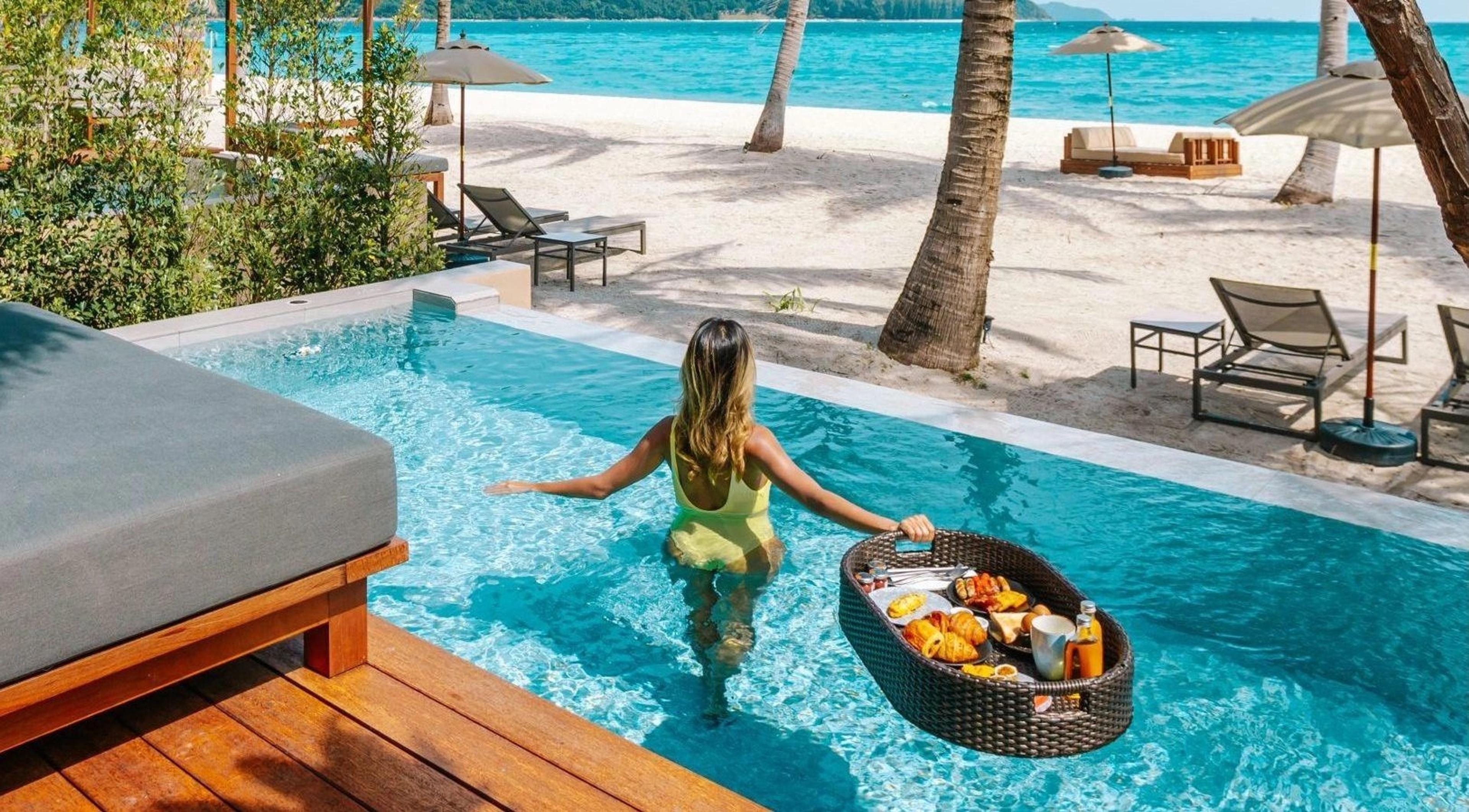a woman wading away from camera through a pool carrying a floating breakfast tray, with sunbeds and the beach behind her