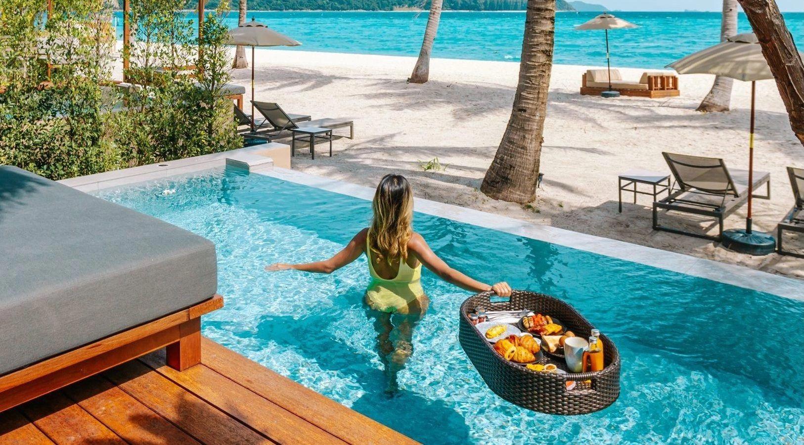 a woman wading away from camera through a pool carrying a floating breakfast tray, with sunbeds and the beach behind her
