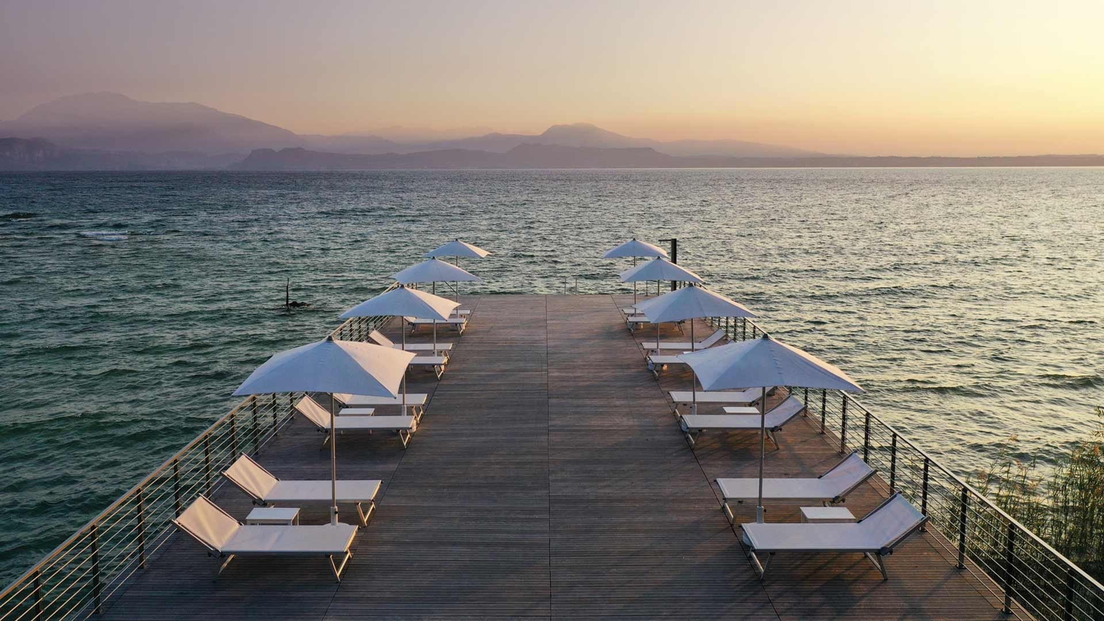 A wooden pier over a lake lined with white sun loungers and umbrellas at sunset, with mountains on the horizon.