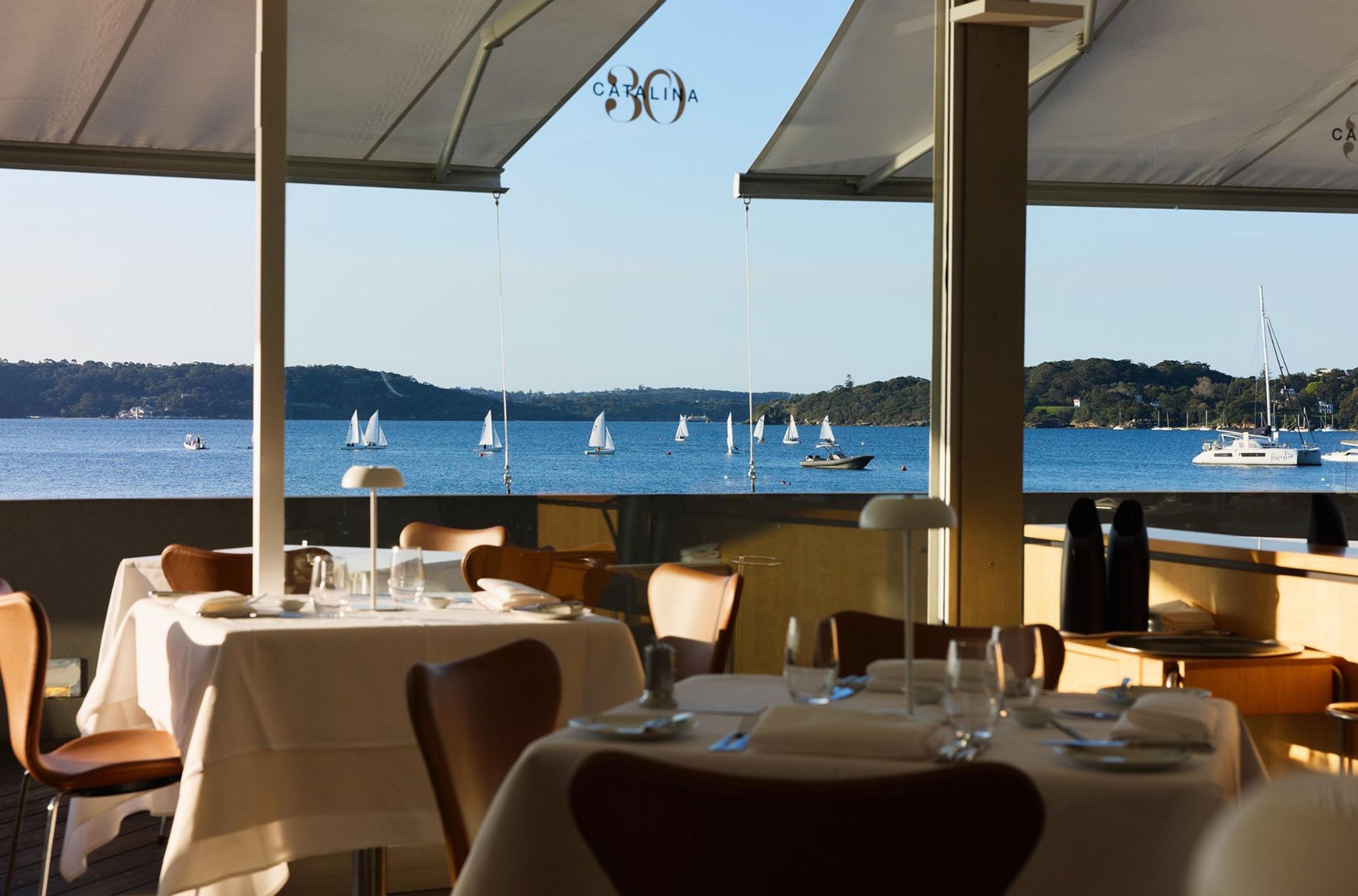 White-clothed tables along a curved wall of glass, with Rose Bay's water and moored boats beyond