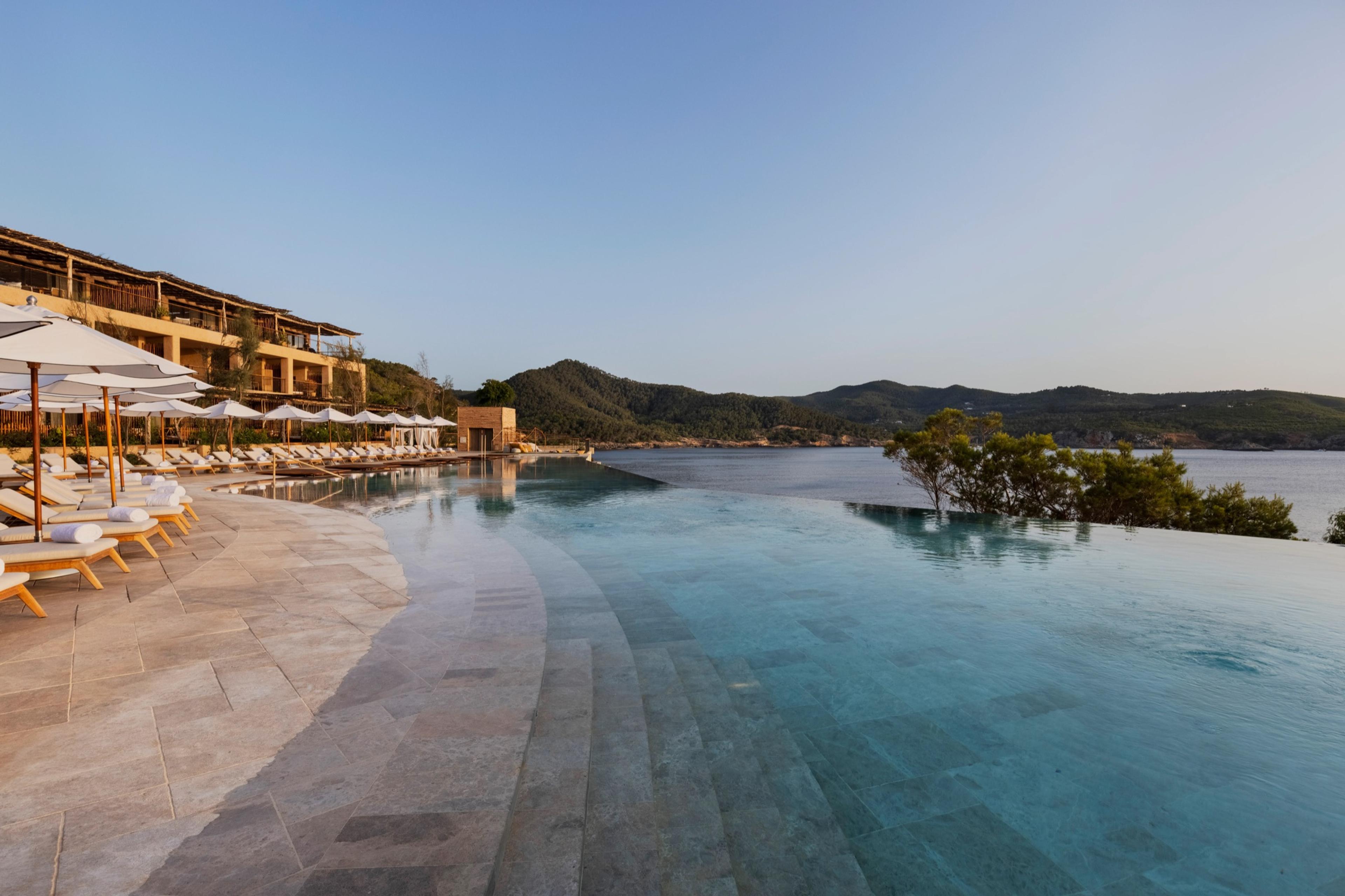 An infinity pool in low evening light, with loungers and parasols on the stone terrace to the left, a low timber-roofed building behind, and calm sea and wooded hills beyond.