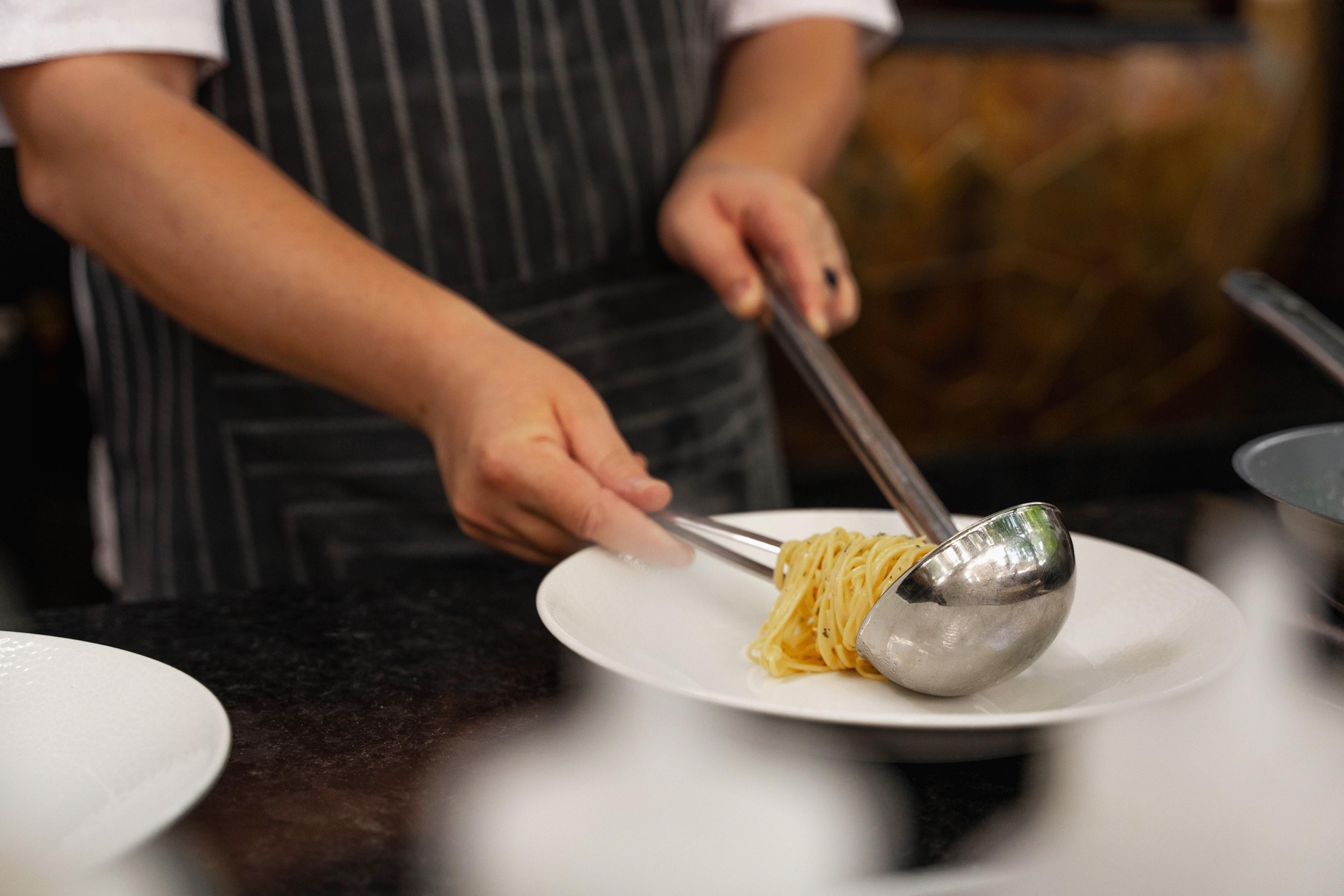 A chef's hands lift a tangle of pasta with tongs and a ladle onto a white plate.