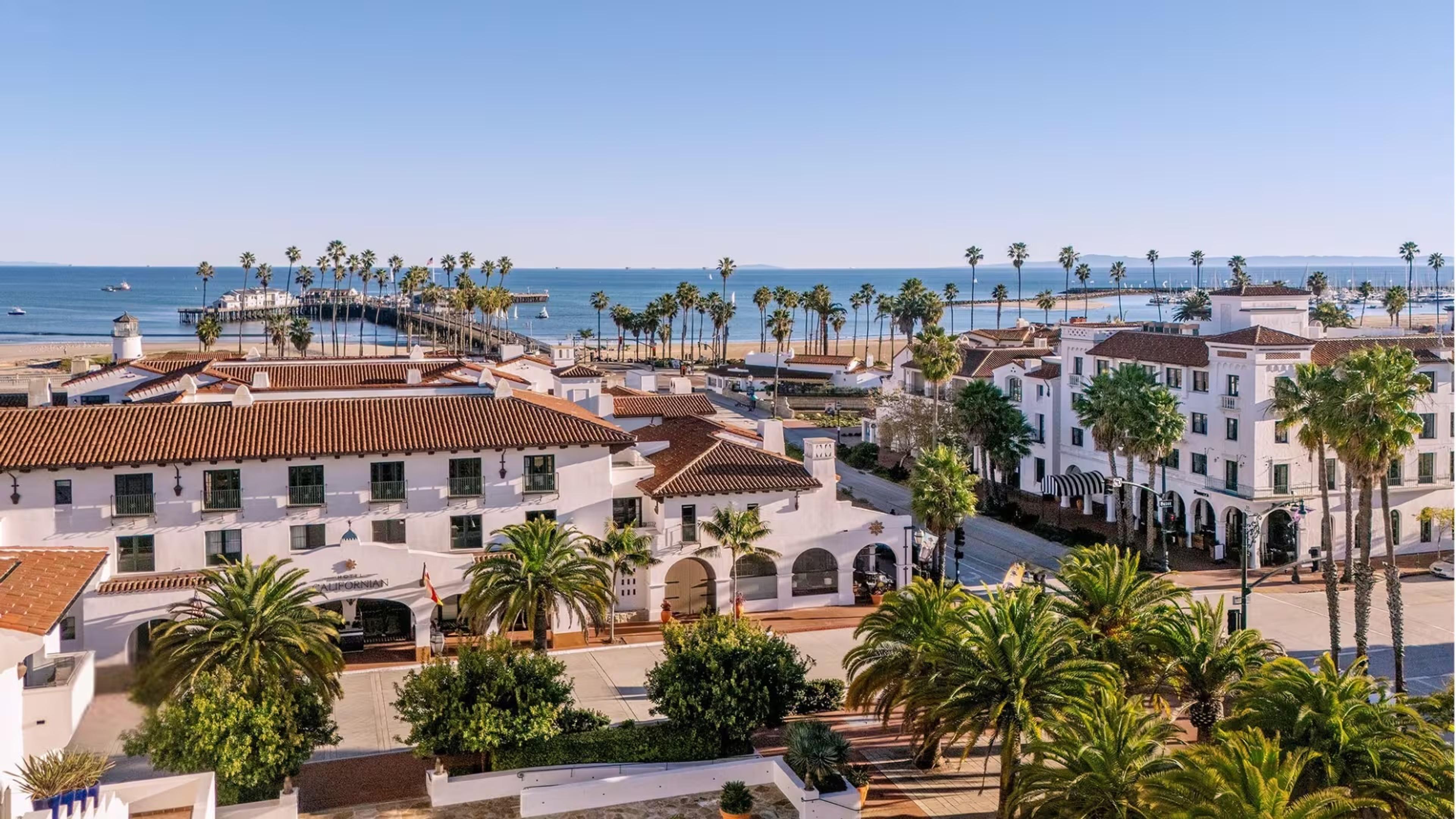 An aerial daytime view of a white Spanish-colonial hotel complex beside a palm-lined beach, a pier, and the ocean, with sailboats and a distant marina.